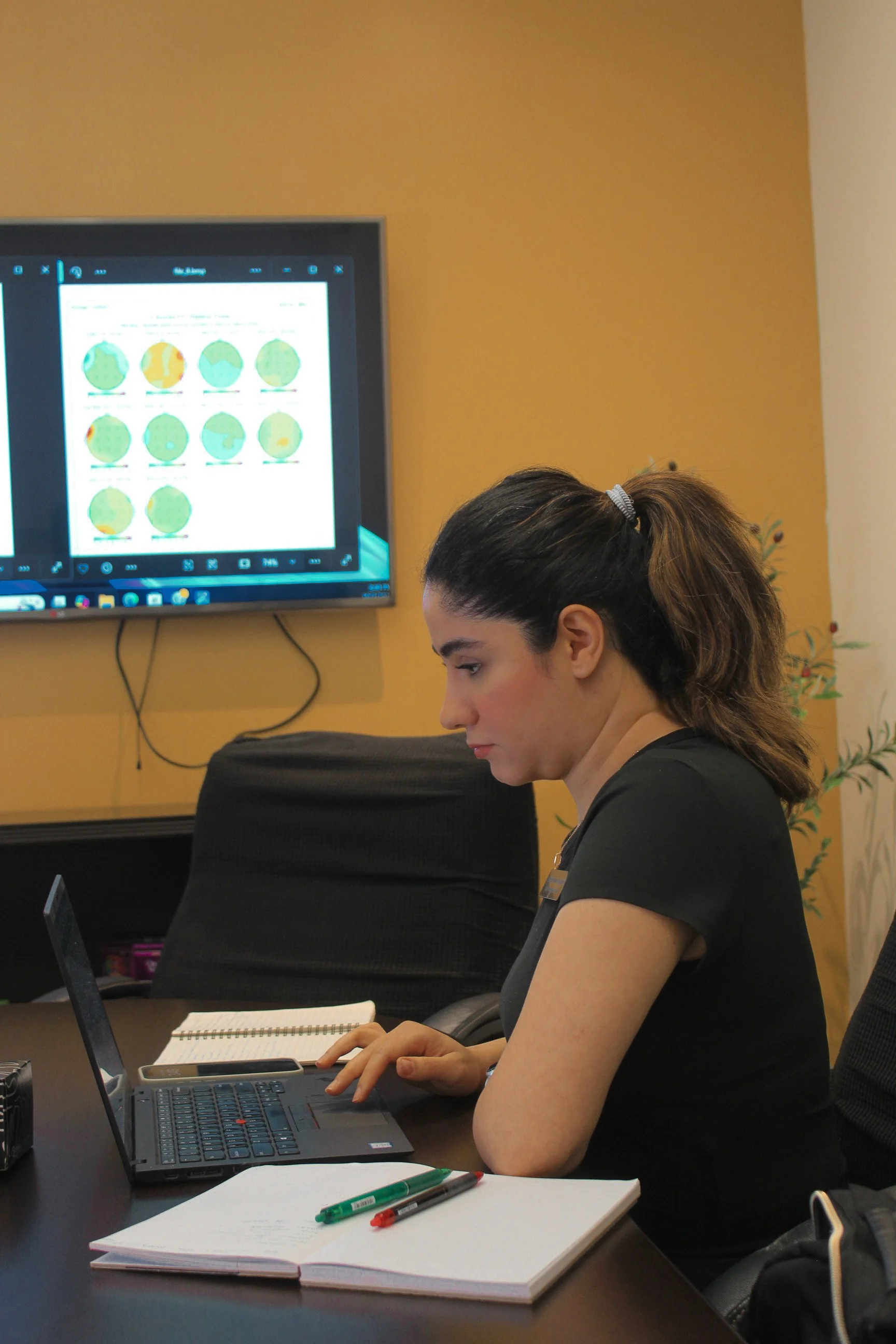 A woman with dark hair tied in a ponytail sits at a conference table working on a laptop, with notebooks and pens in front of her. She is looking at her laptop screen with a large monitor behind her displaying a colorful chart or infographic.