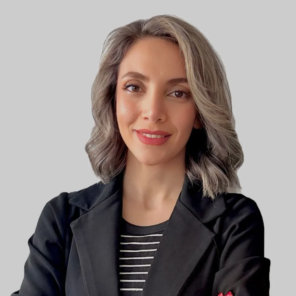 A woman with shoulder-length wavy hair, light skin, and makeup, smiling at the camera, wearing a black jacket over a black and white striped shirt, standing against a plain light gray background.
