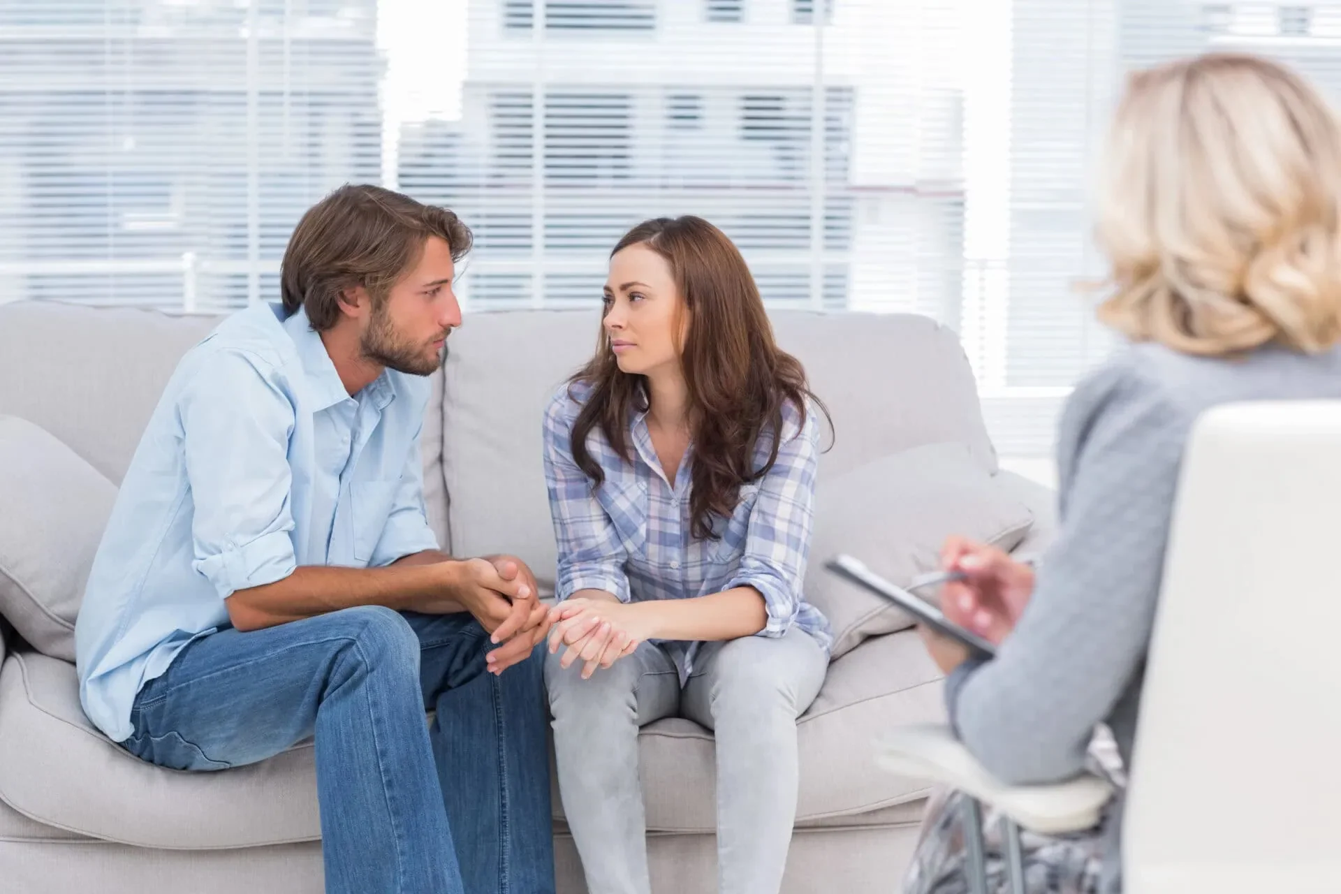 A man and woman sitting on a couch holding hands, talking seriously with a therapist taking notes.