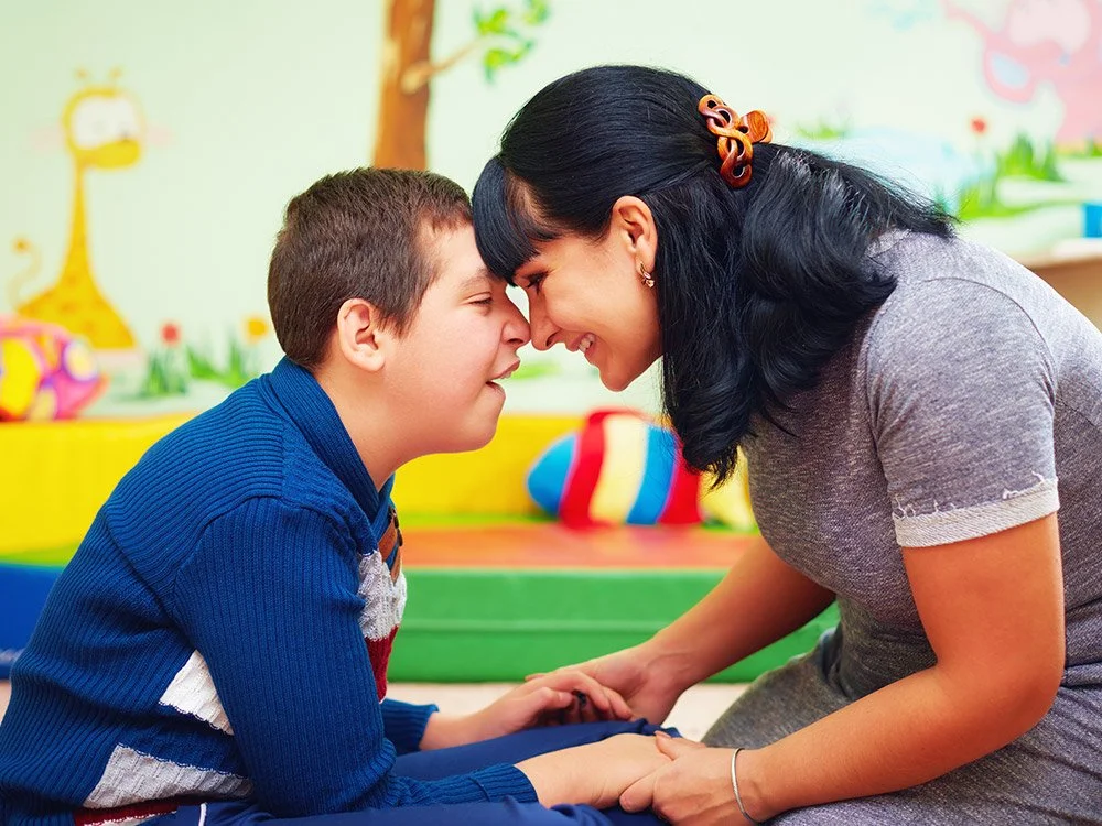 A woman and a boy touching foreheads and smiling at each other in a colorful playroom with toys and mural paintings.
