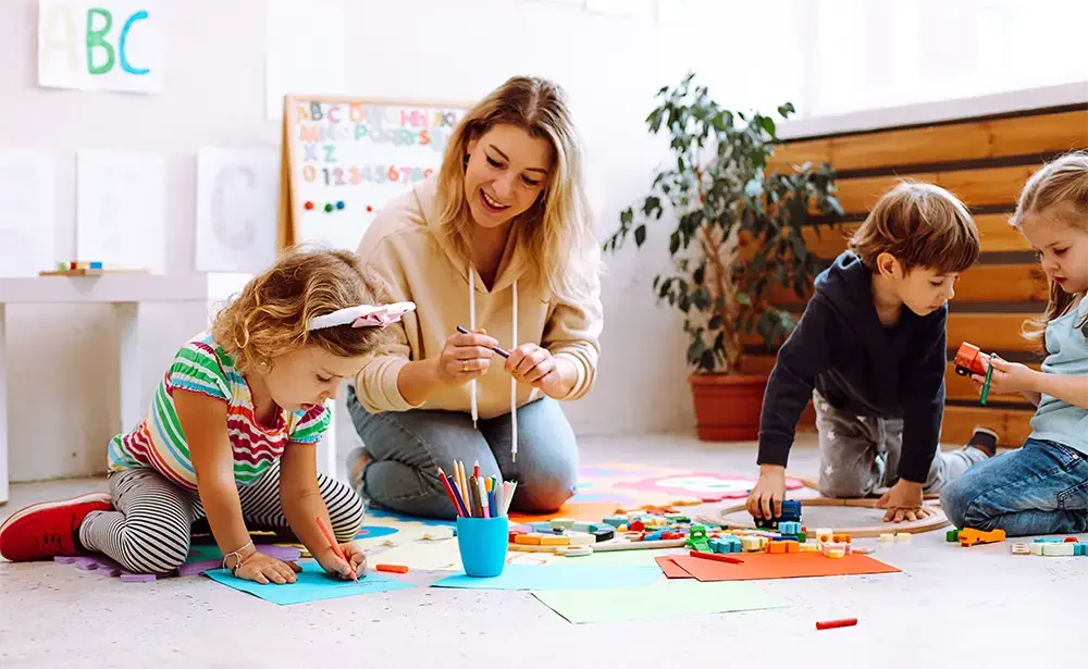 Teacher and three children drawing and playing with colorful paper and toys on a classroom floor.