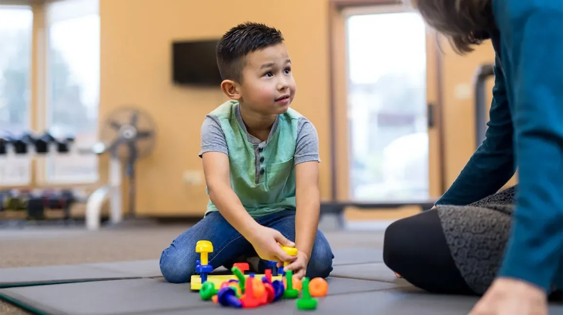 Young boy sitting on a mat playing with colorful plastic toy pieces, adult nearby, indoor setting.