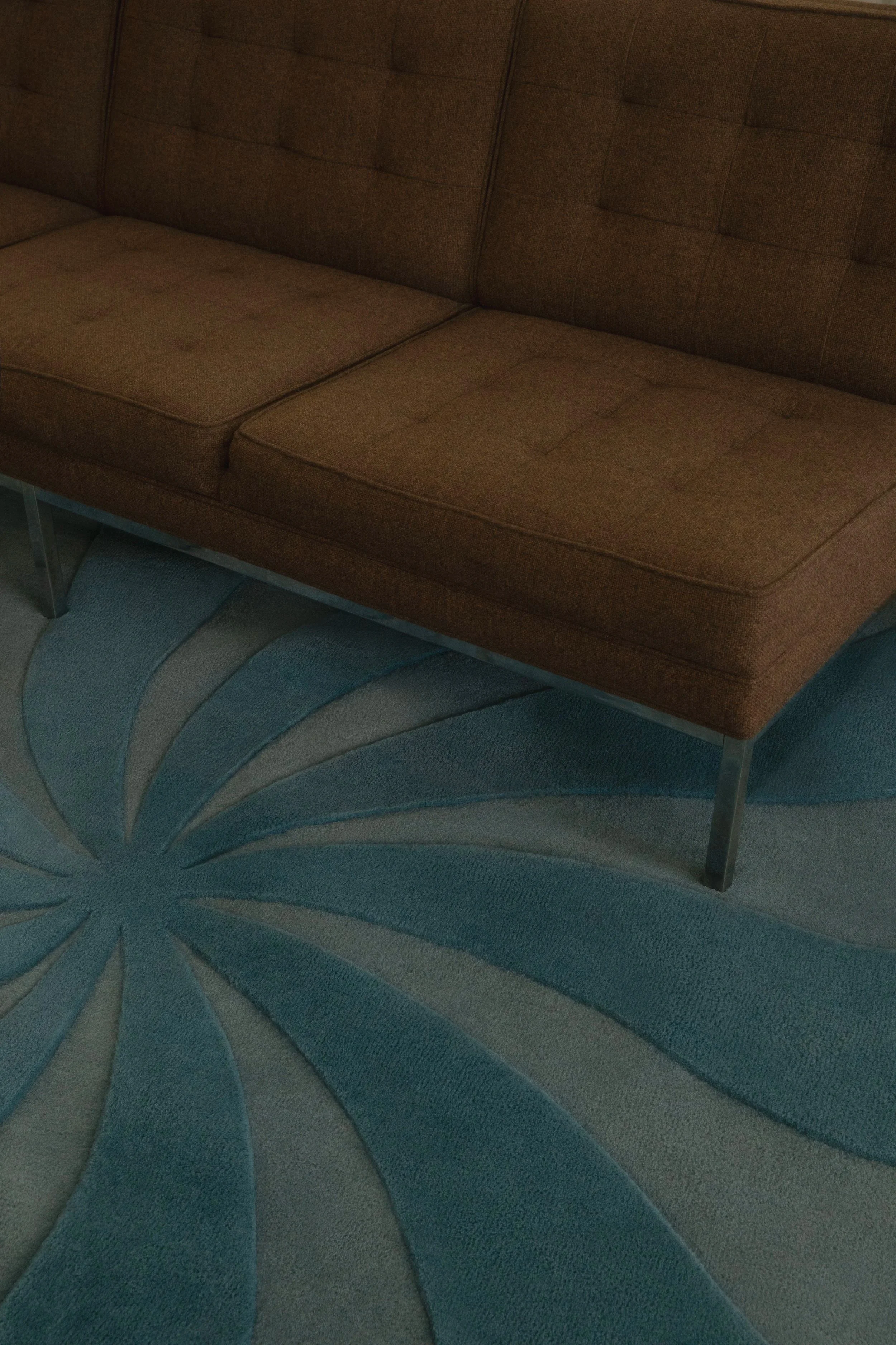 Close-up of a mid-century–style brown upholstered sofa with tufted cushions and slender metal legs, positioned on a teal and gray rug featuring a radiating, starburst-style pattern.