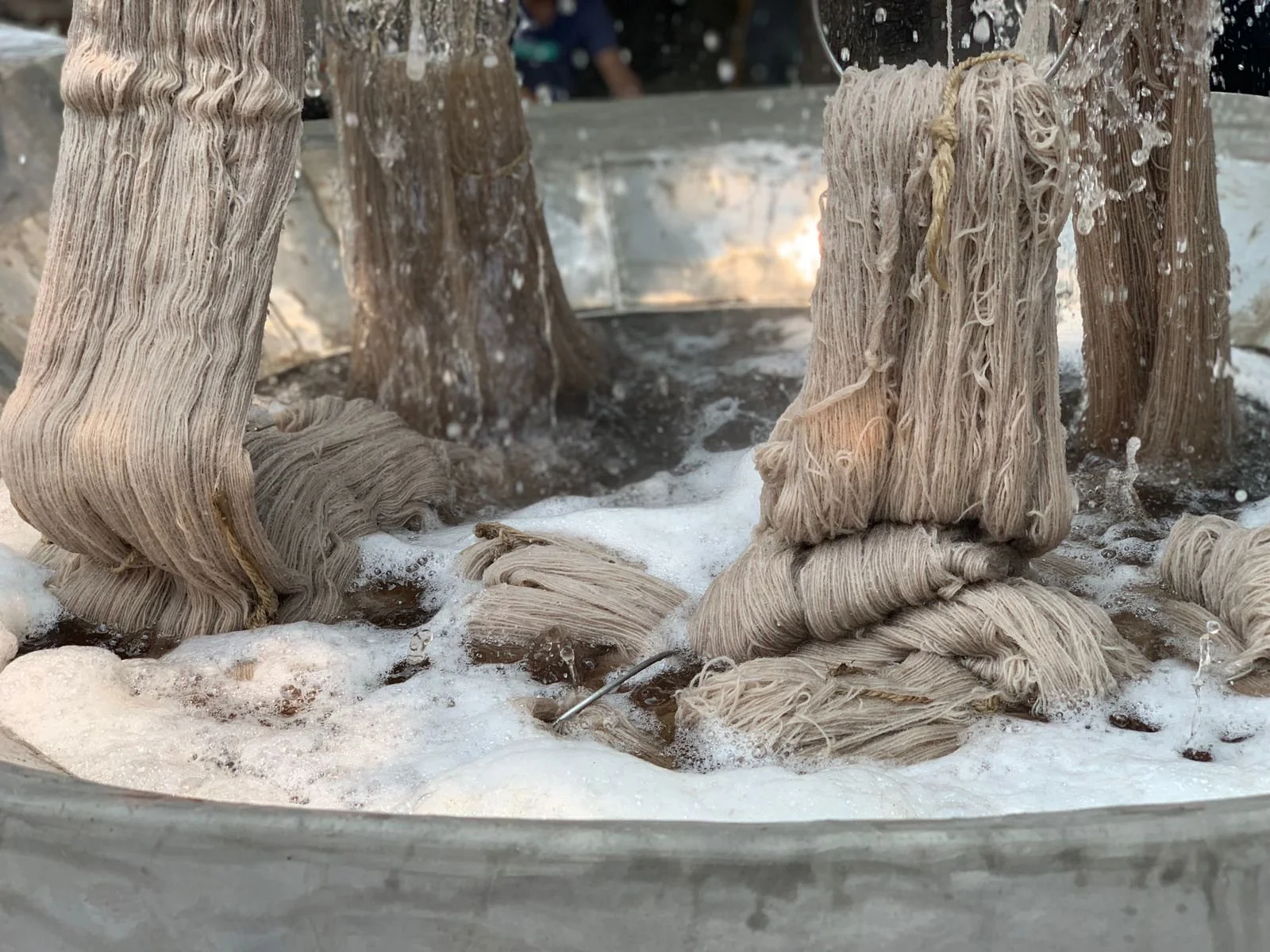 Close-up of traditional hand-pulled noodle drying process, with wet, tangled noodles hanging from vertical rods in a large metal basin containing water and soap, outdoors.