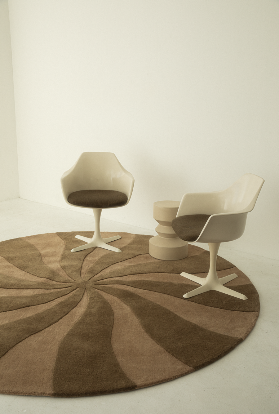 Minimalist interior with two modern white chairs with brown cushioned seats, a round side table, and a large patterned brown rug on a white floor and wall.