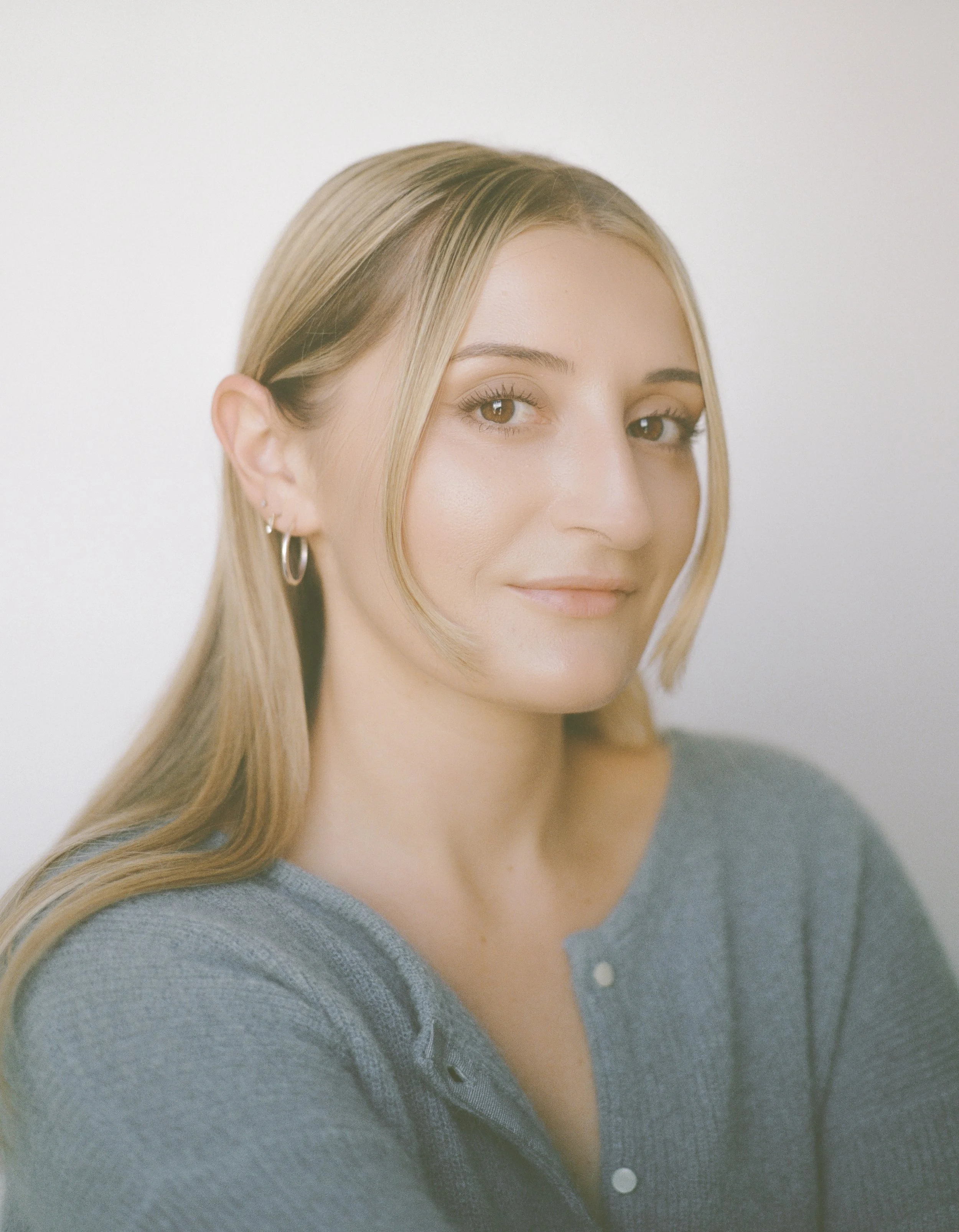 A young woman with blonde hair and light makeup, wearing a gray top and small hoop earrings, looking at the camera with a slight smile.