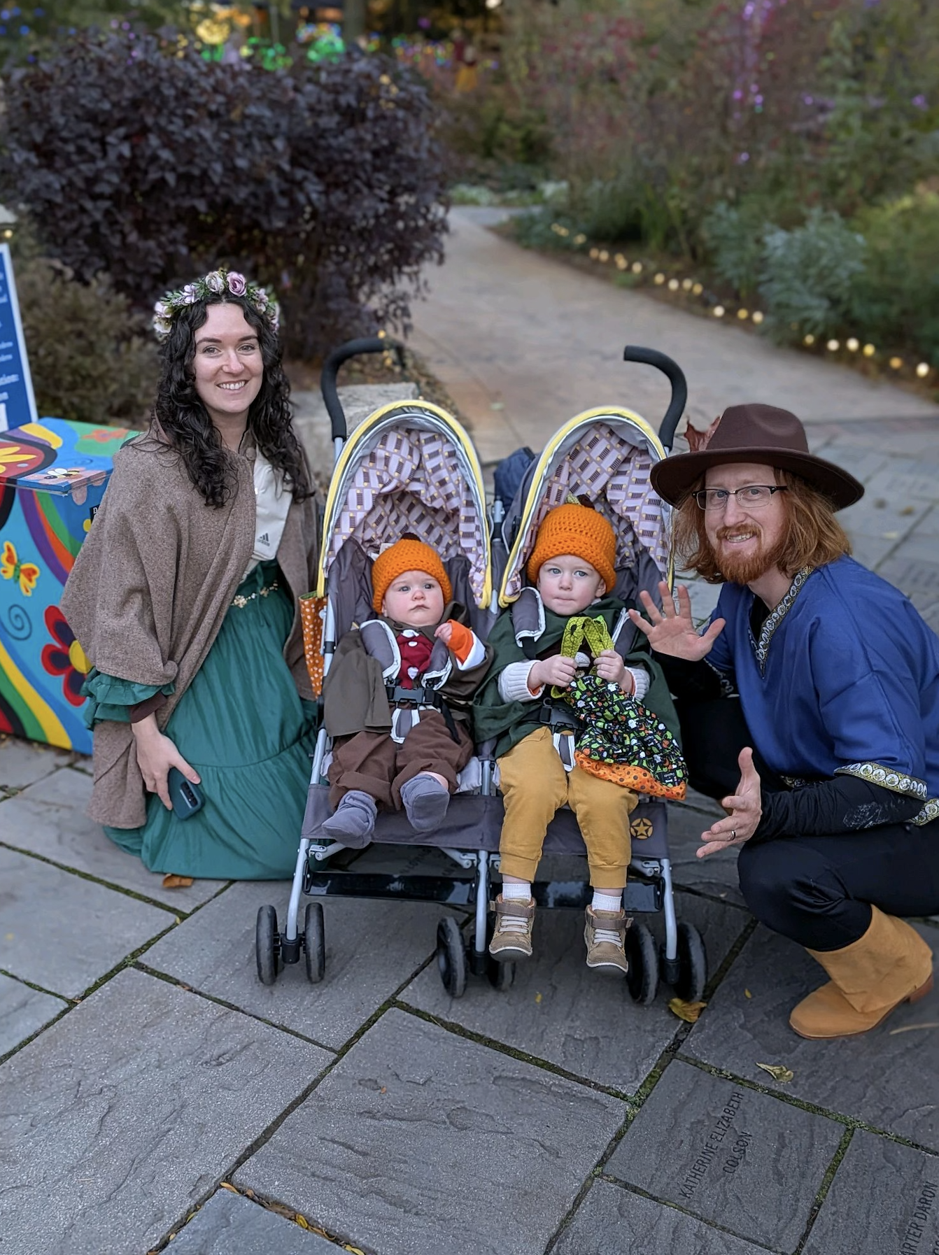 A woman and a man are outside, kneeling next to two children in stroller seats. The woman is wearing a floral crown, a brown shawl, and a green skirt, and is holding a phone. The man is wearing a brown wide-brimmed hat, glasses, a blue shirt, and black pants. The children are dressed in warm clothes with orange knit hats. The scene appears to be during the evening in a park or garden with plants, a pathway, and lights in the background.