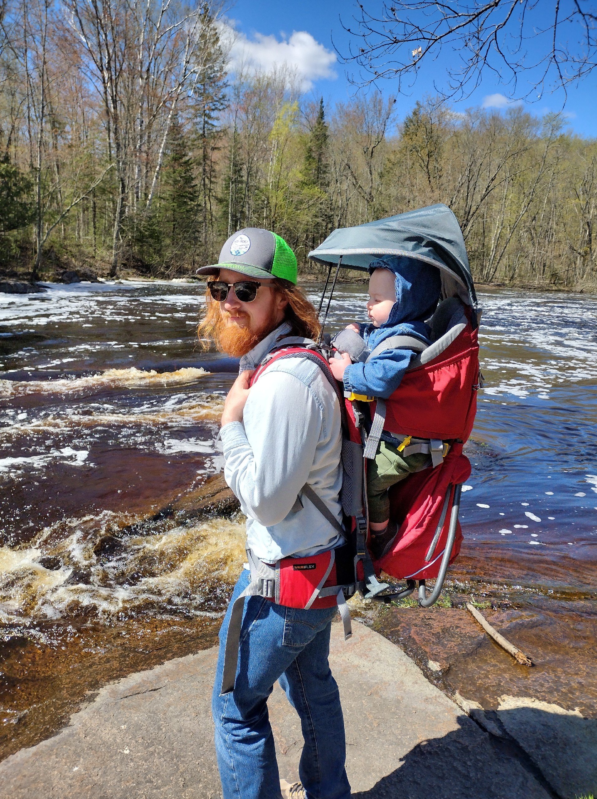 Man carrying a young child in a red backpack carrier by a river with trees and blue sky in the background.