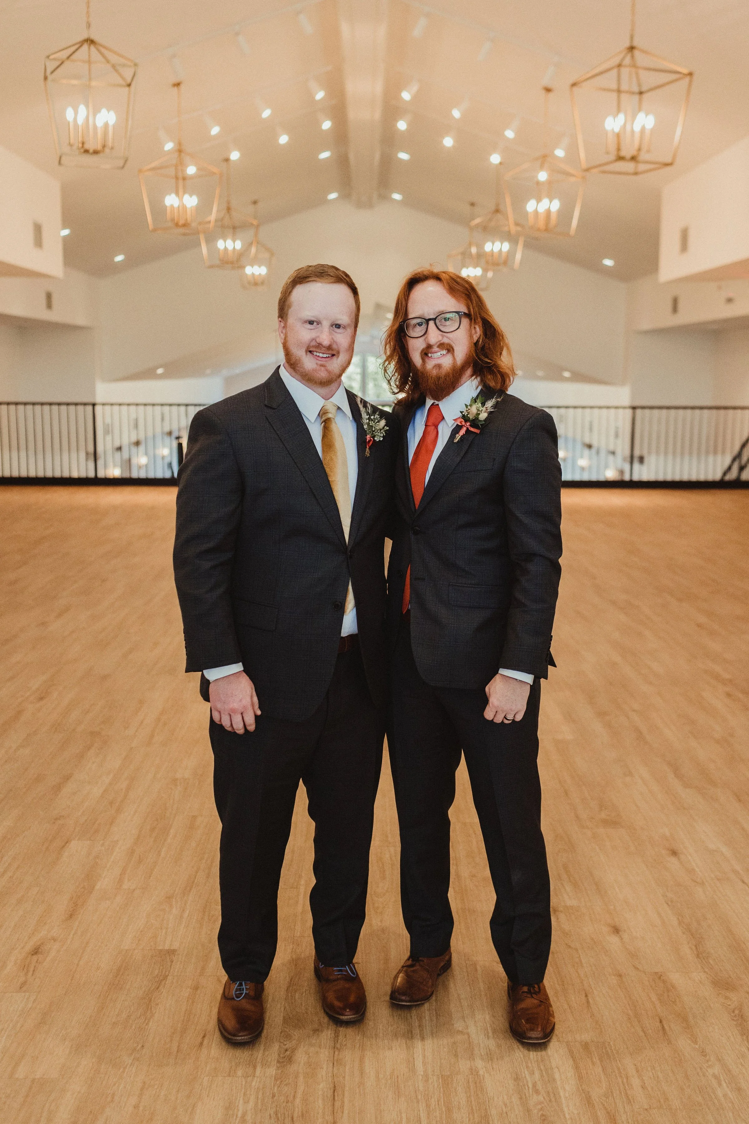 Two men in suits standing in a large, well-lit indoor space with wooden flooring and decorative hanging lights.