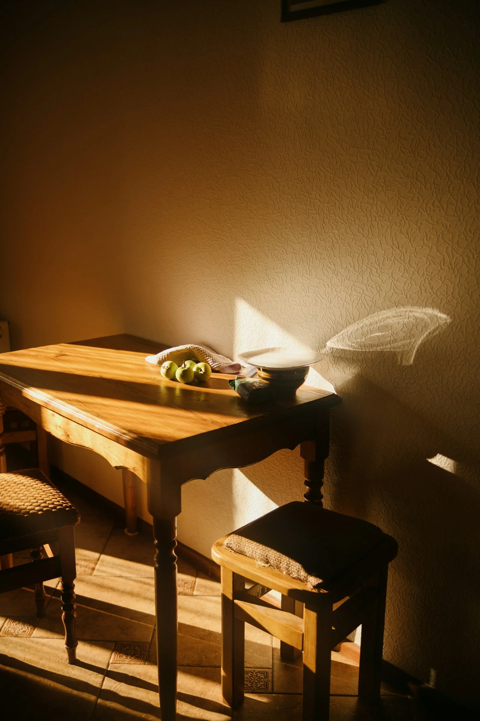 Sunlight illuminating a small wooden dining table with three green apples, a beige cloth, and stacked bowls, casting shadows on the textured cream wall and tiled floor.