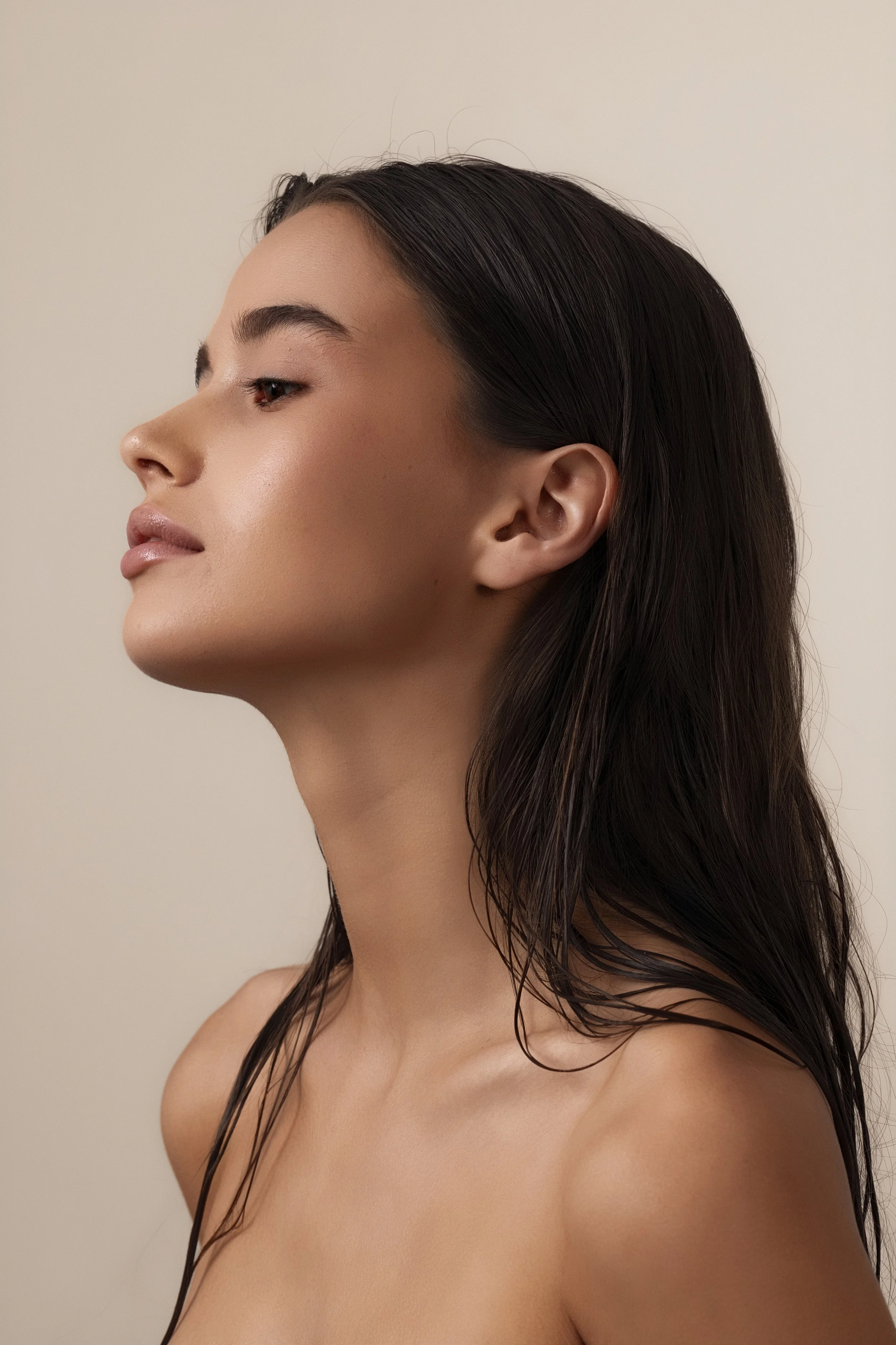 A young woman with long dark hair and smooth skin, looking to the side, set against a plain neutral background.