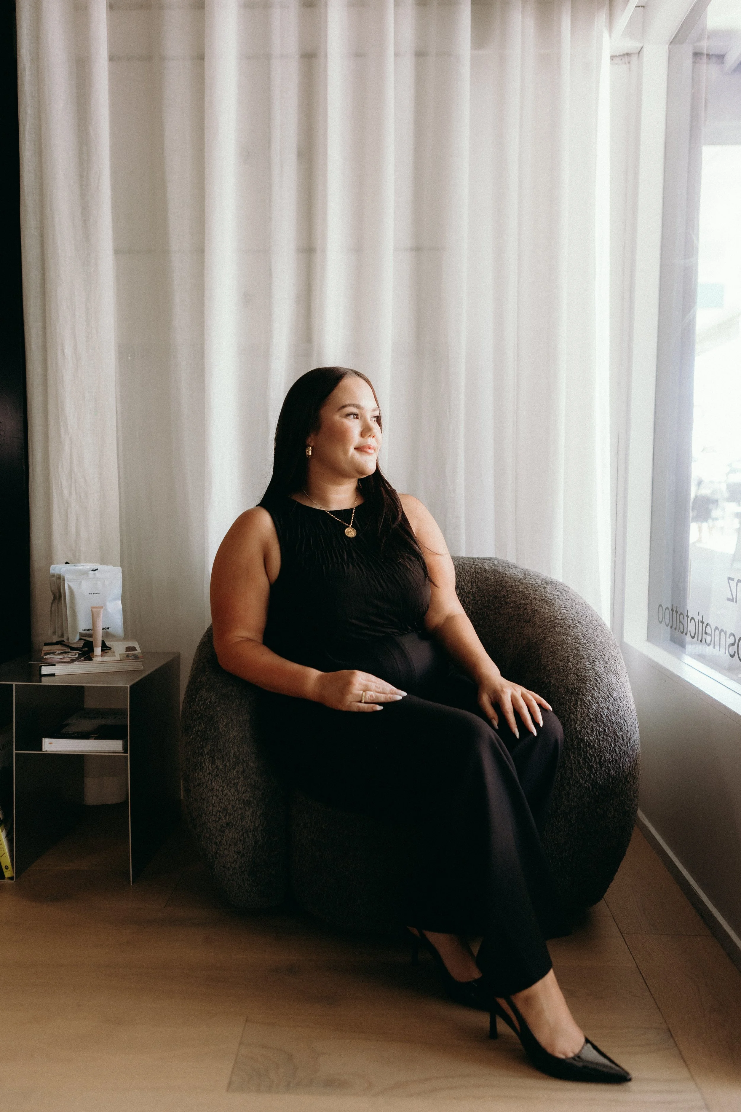 A woman sitting on a gray armchair by a window with white curtains, wearing a black dress and high heels, looking to the side smiling.