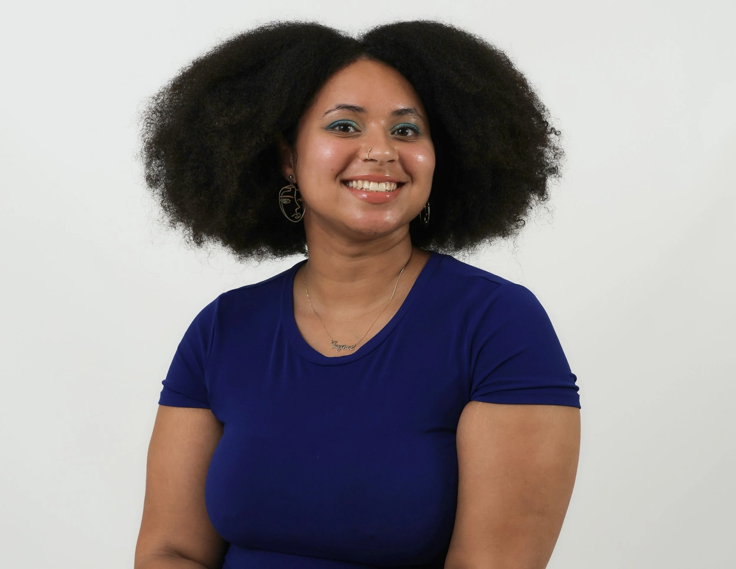 A light skinned Black woman with curly black hair, wearing blue eyeshadow, earrings, a nose ring, a necklace that says Tayoncé and a blue shirt, smiling in front of a plain white background.