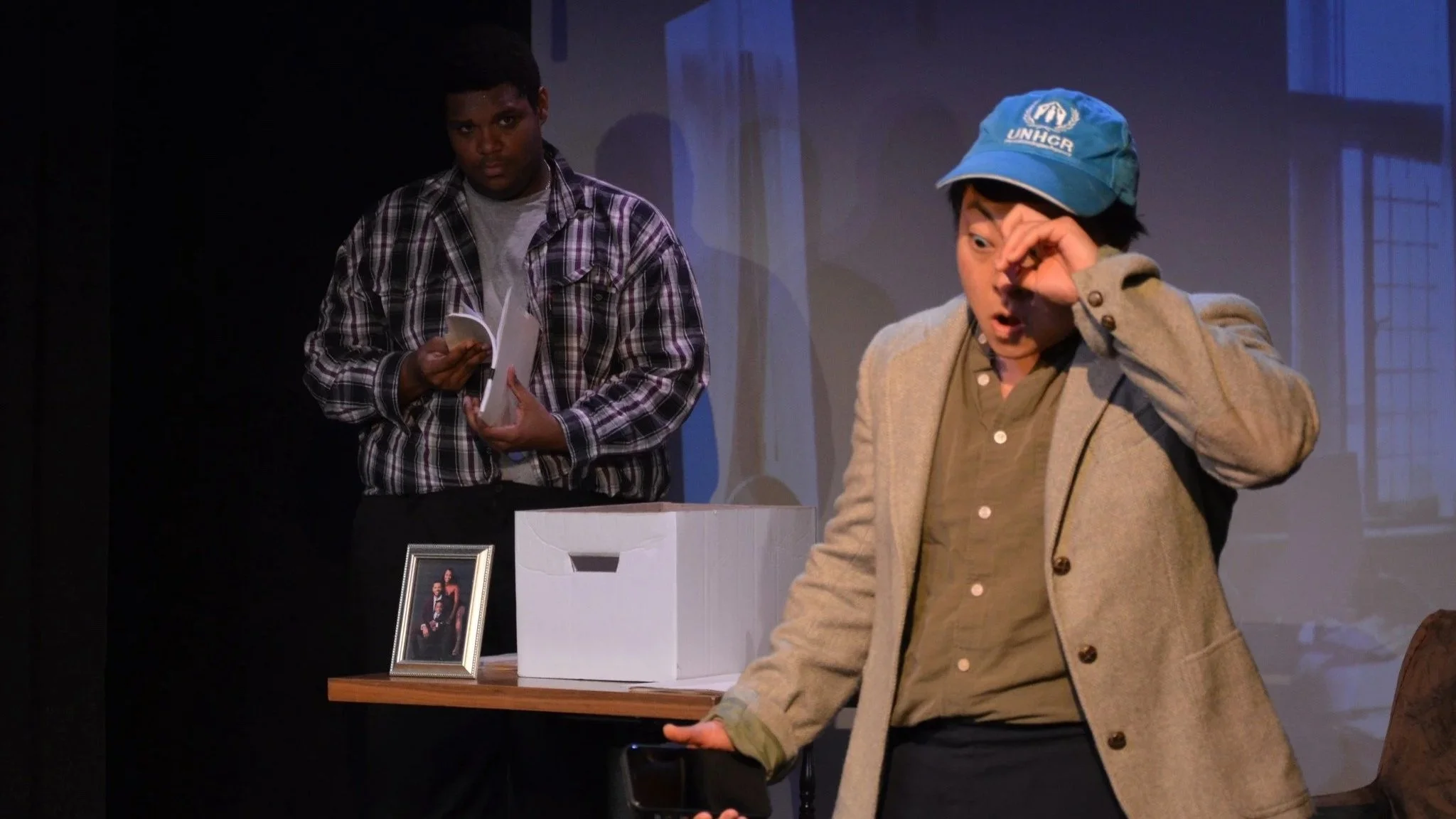 Two masculine people on a stage, one in a plaid shirt holding papers and the other in a beige coat and blue UNHCR cap, with a table displaying a framed photograph and a white box beside it.