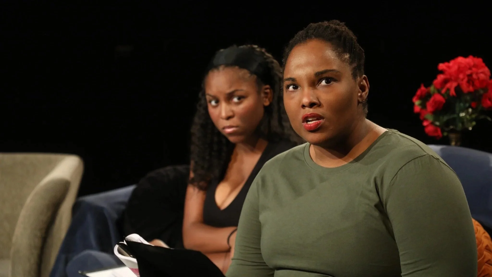 Two women sitting on a blue couch, with red flowers in a pot in the background, with one woman in the foreground having short hair and wearing a green shirt, and the other woman in the background with long, braided hair wearing a black top.