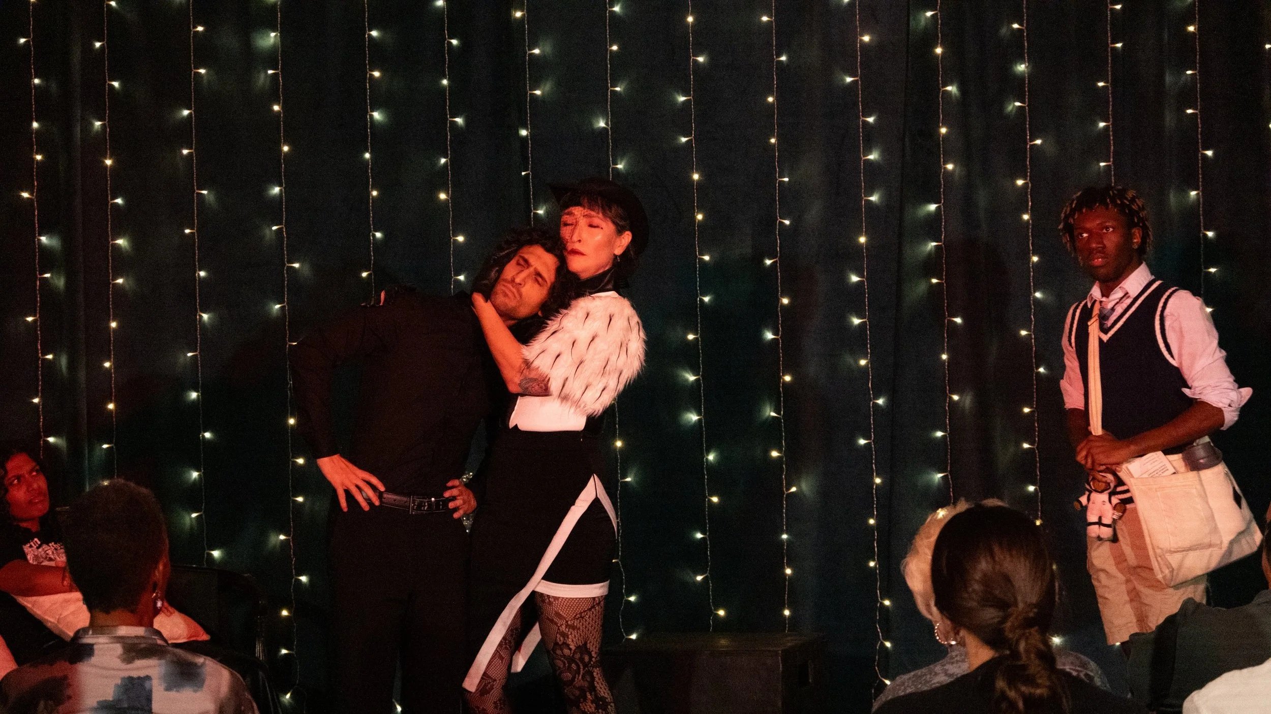 Three people on stage in front of a backdrop of hanging string lights. One person is embracing a woman, both with serious expressions, while a third person watches. Audience members are visible in the foreground.