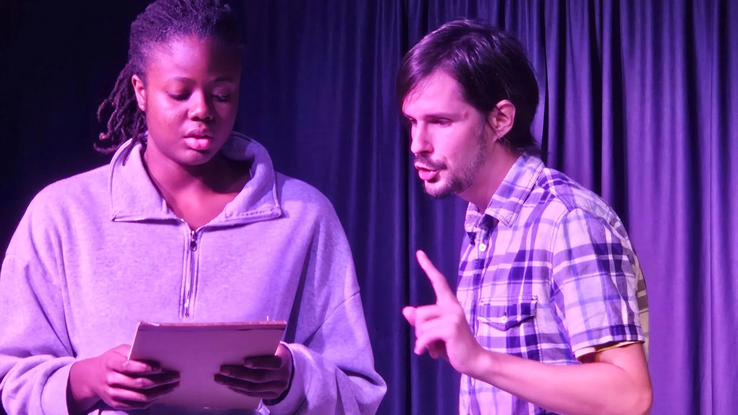 Two young adults having a conversation on stage, one holding a script and the other gesturing with their hand, in front of a purple curtain.