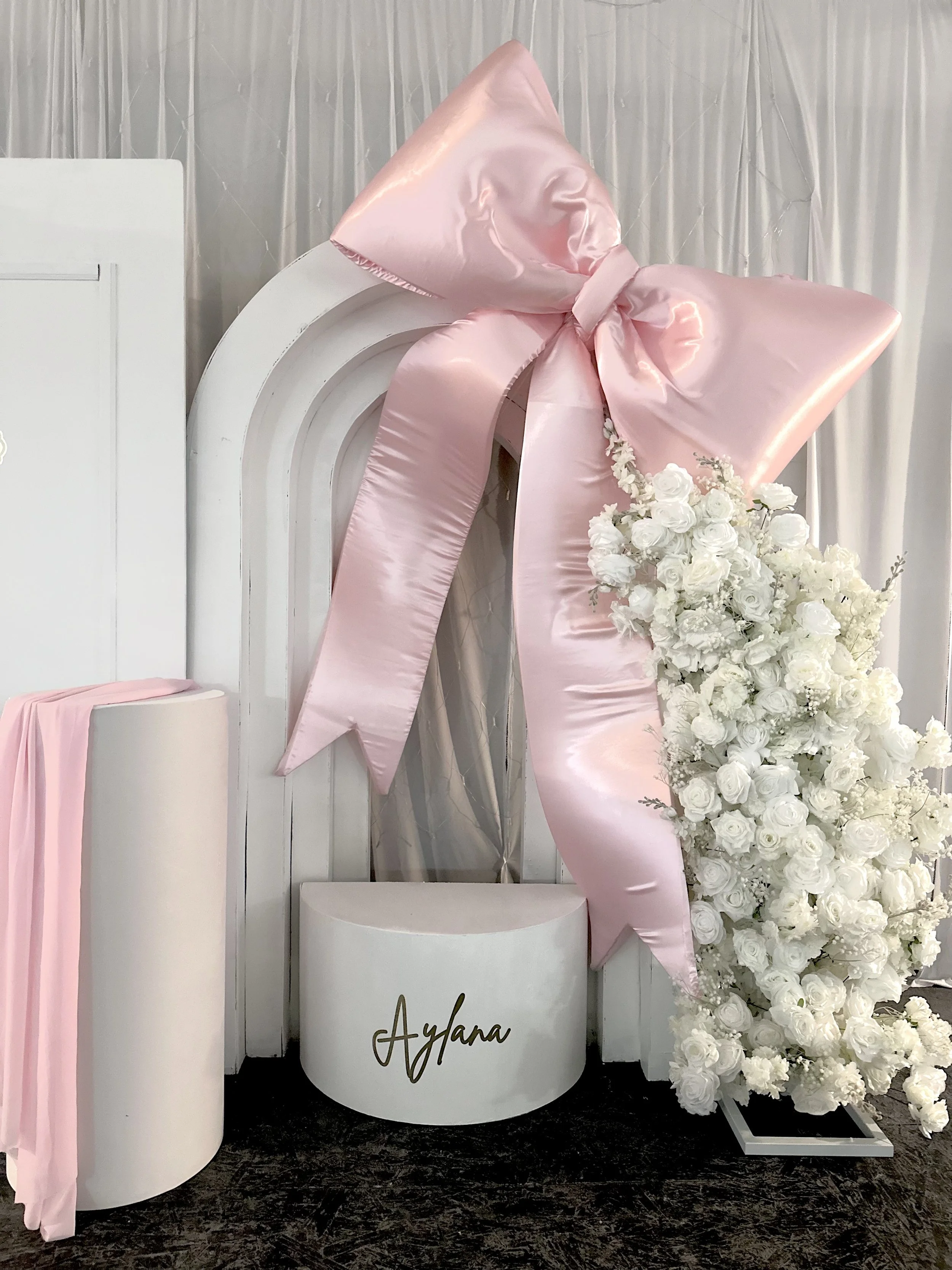 White circular display with the word "Alyana" written in gold, pink satin fabric bow, and a large arrangement of white roses, with a white curtain background.
