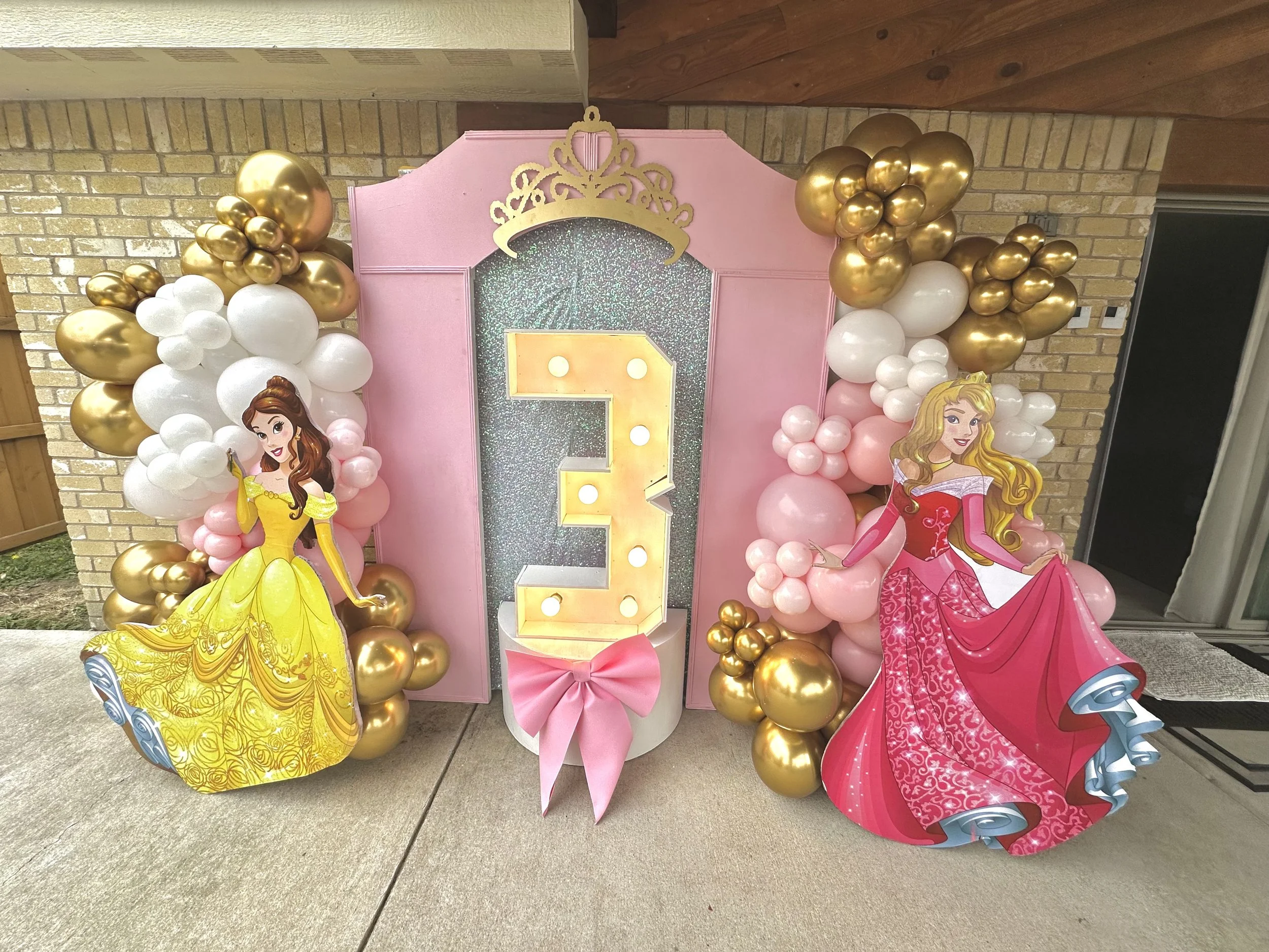Pink princess-themed birthday backdrop decorated with gold and white balloons, featuring cutouts of Disney princess characters Belle and Aurora, and a large illuminated number one in the center.