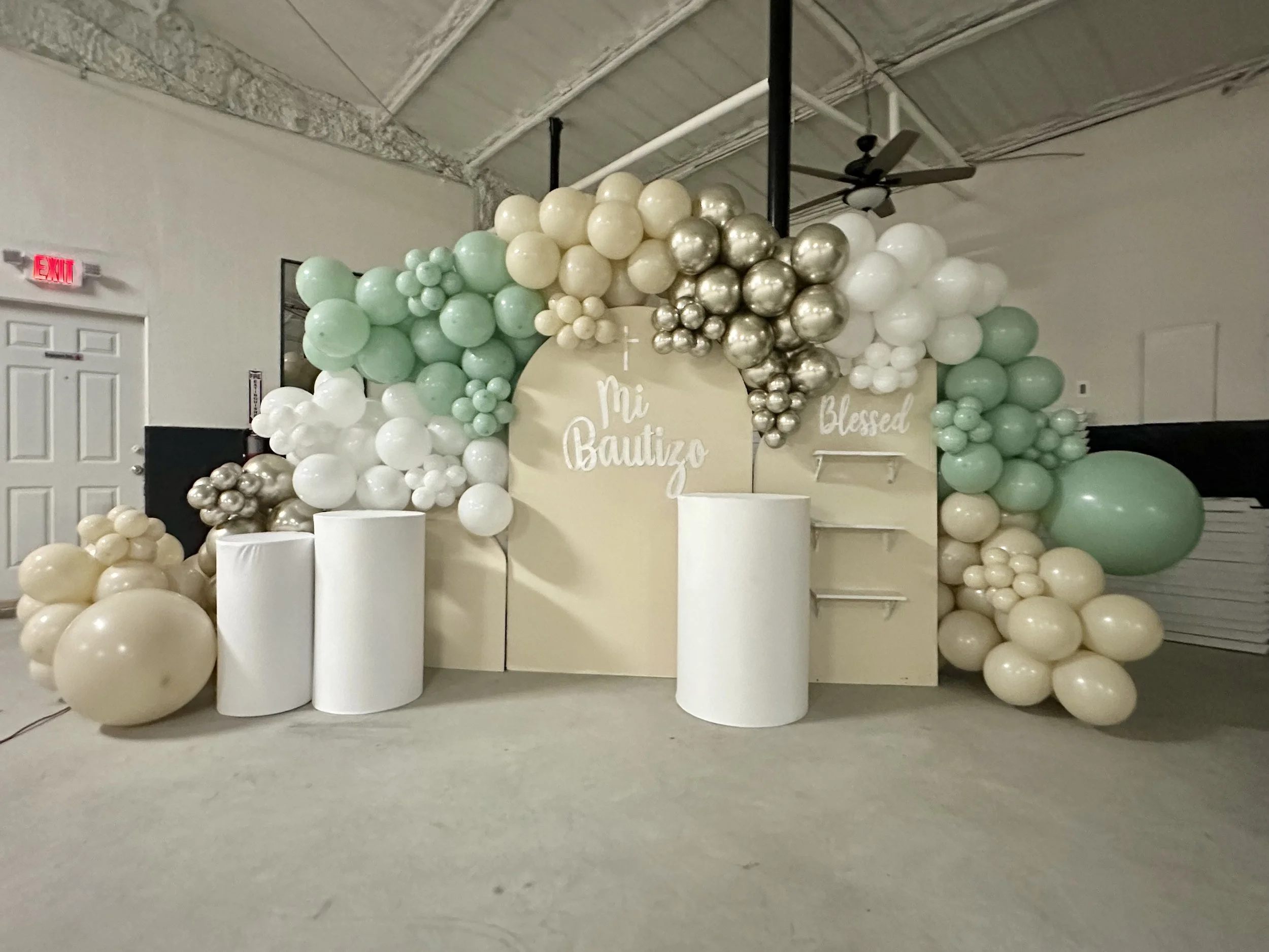 Decorative balloon arrangement with pastel green, white, and metallic silver balloons, with a beige backdrop that has the words 'My Baptize' and 'Blessed' written on it, set in an indoor space with a ceiling fan and an exit sign visible.
