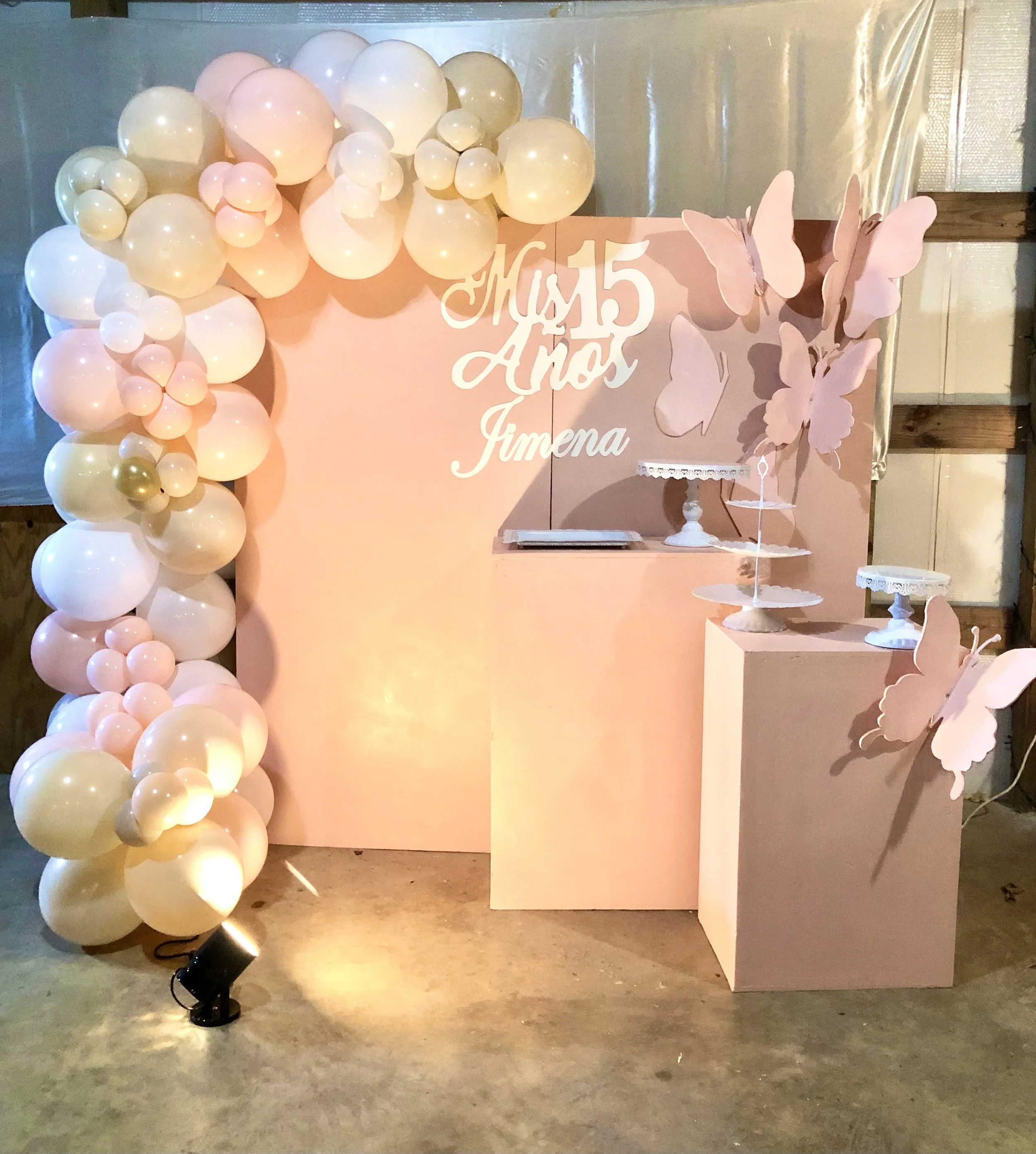 Decorative backdrop with cream, peach, and gold balloons, a pink sign reading 'Mãe 15 Amor Fimena' with butterfly and floral cutouts, and star-themed tables and chairs, indicating a celebration, possibly a birthday or special event.