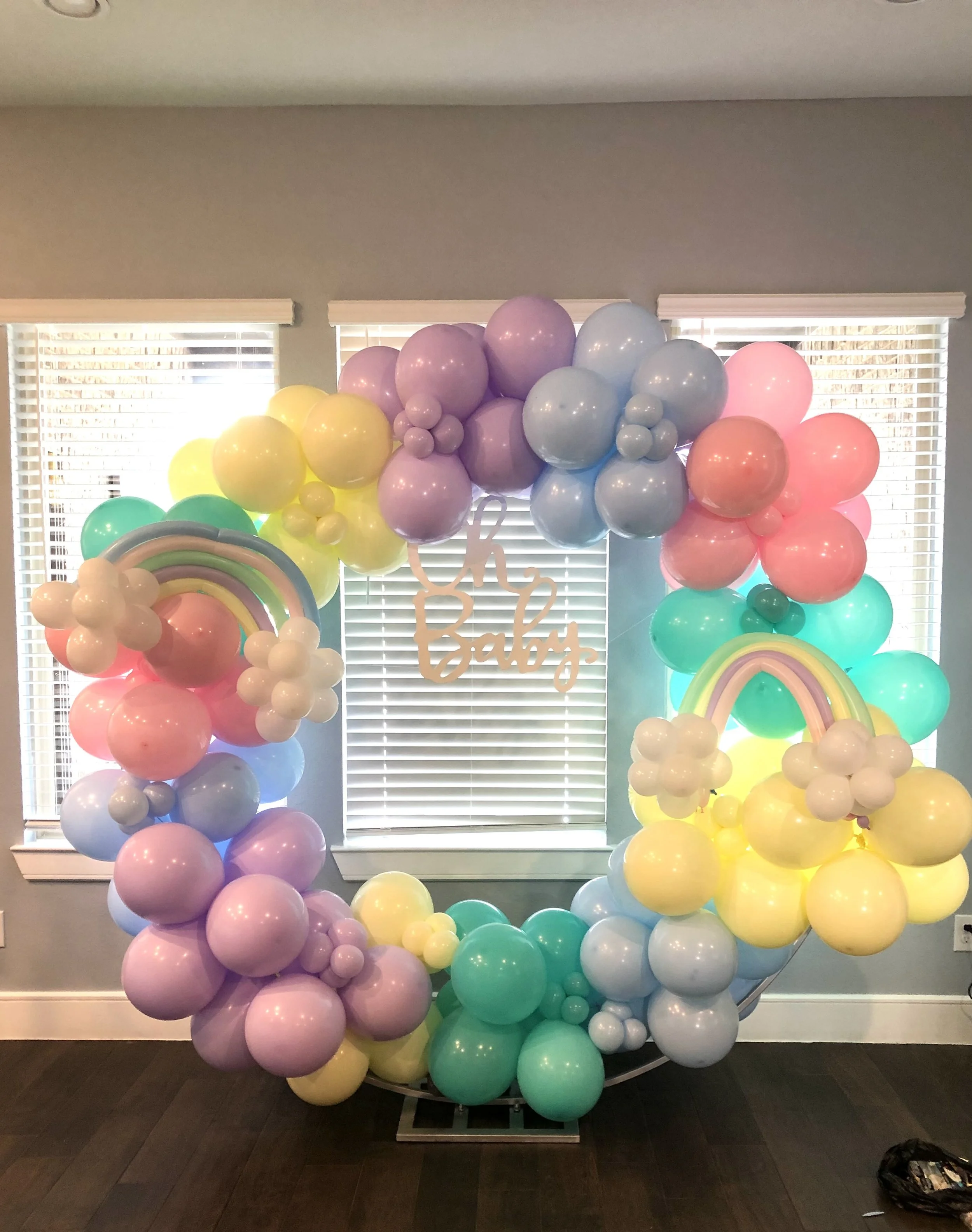 Colorful balloon arrangement with pastel pink, purple, yellow, green, and blue balloons, including large flower-shaped balloons, with a sign that says 'oh Baby' tied to the balloons, set up indoors in front of a window with blinds.