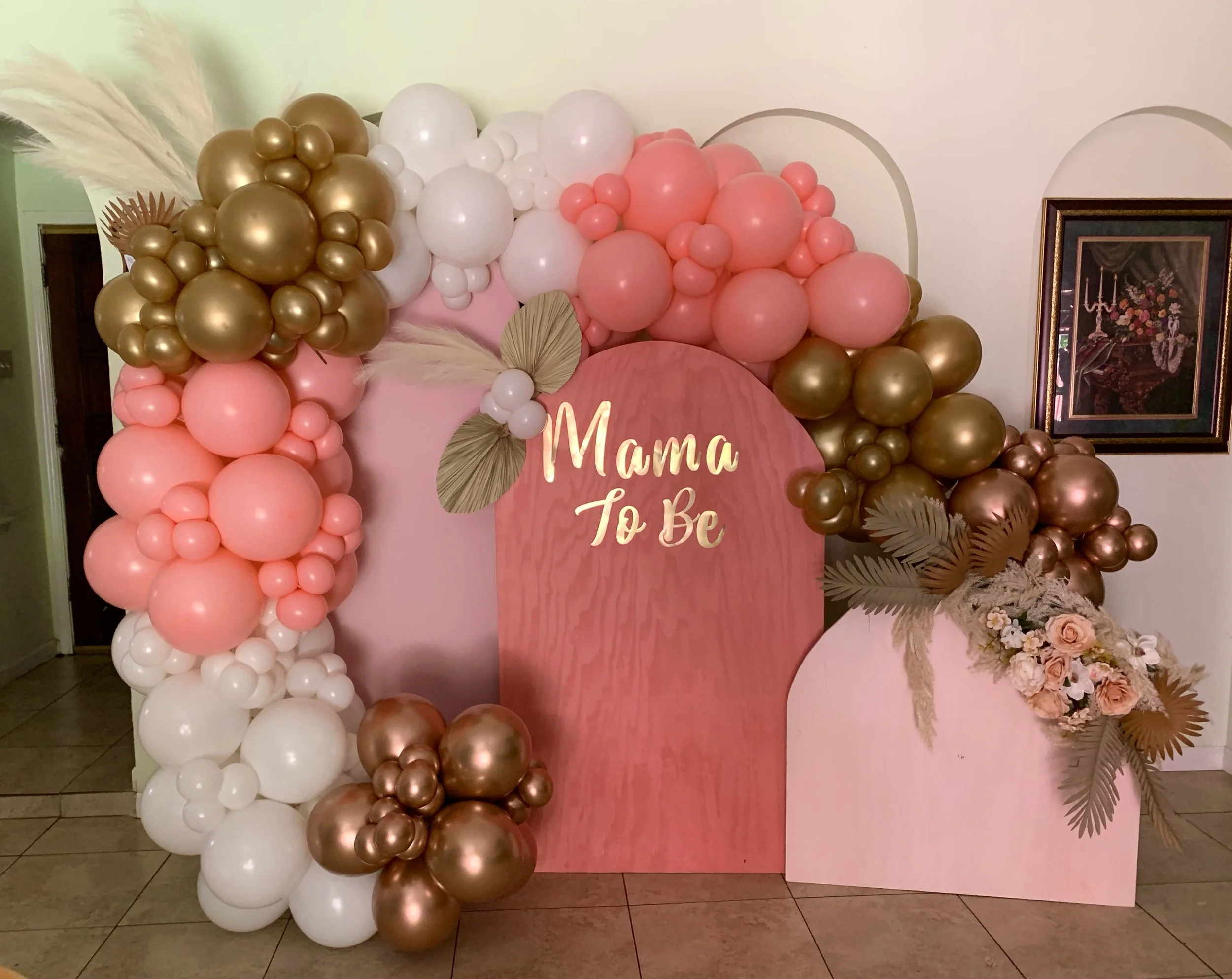 Balloon display with pink, white, and gold balloons arranged on a pink backdrop, with a sign that reads 'Mama to Be' and floral decorations.