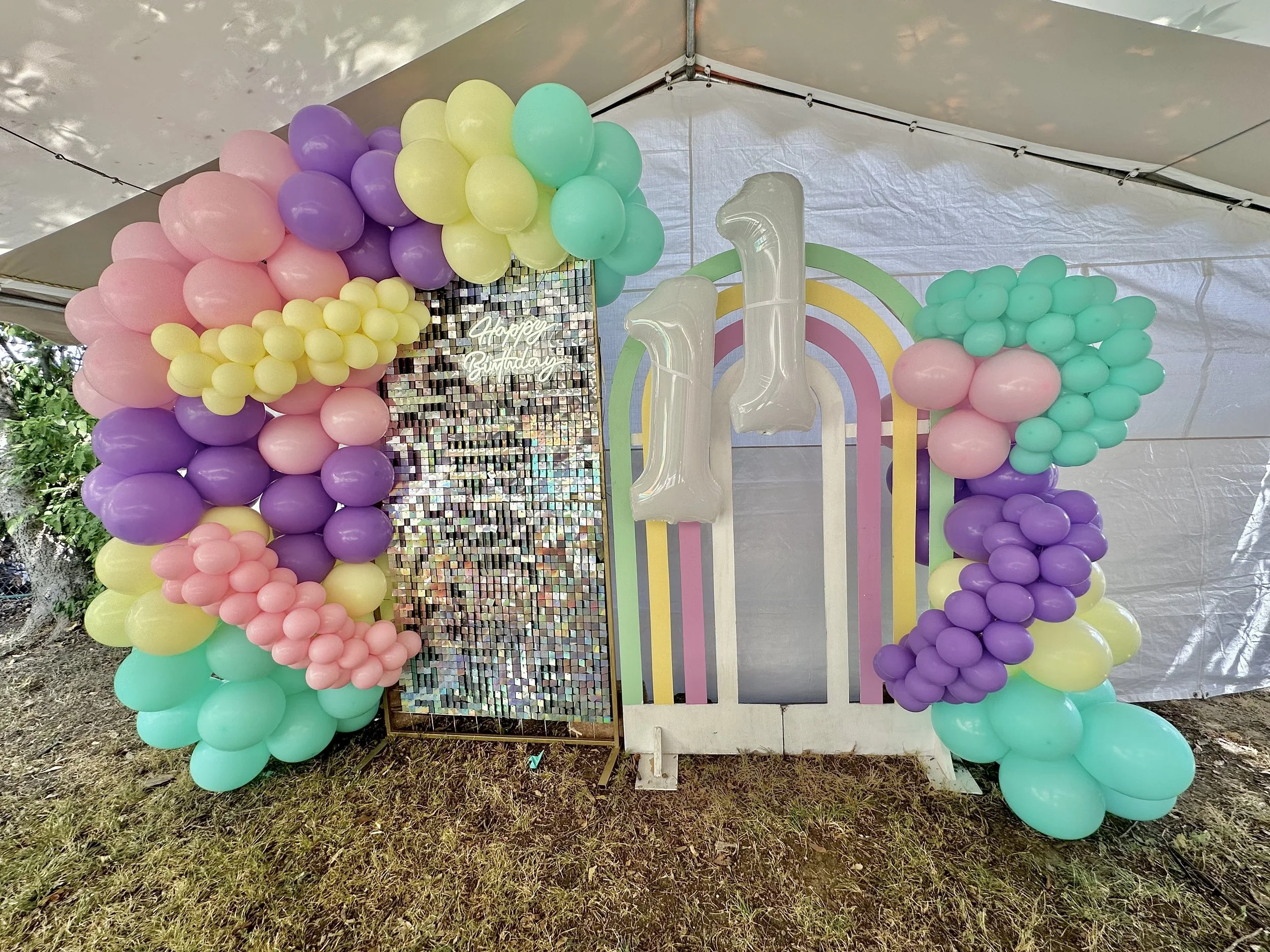 Colorful balloon arch in pastel colors including pink, purple, yellow, and mint green with a backdrop featuring a rainbow and a 'Happy Birthday' sign.