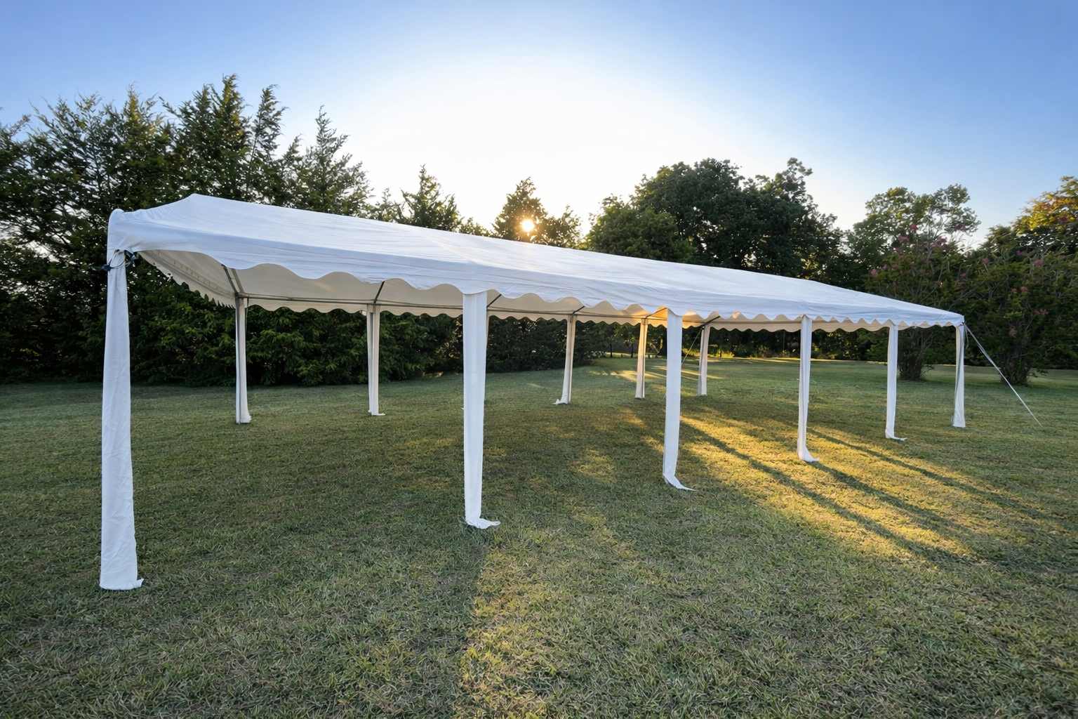 A large white event tent set up on a grassy field with sunlight shining through trees in the background.