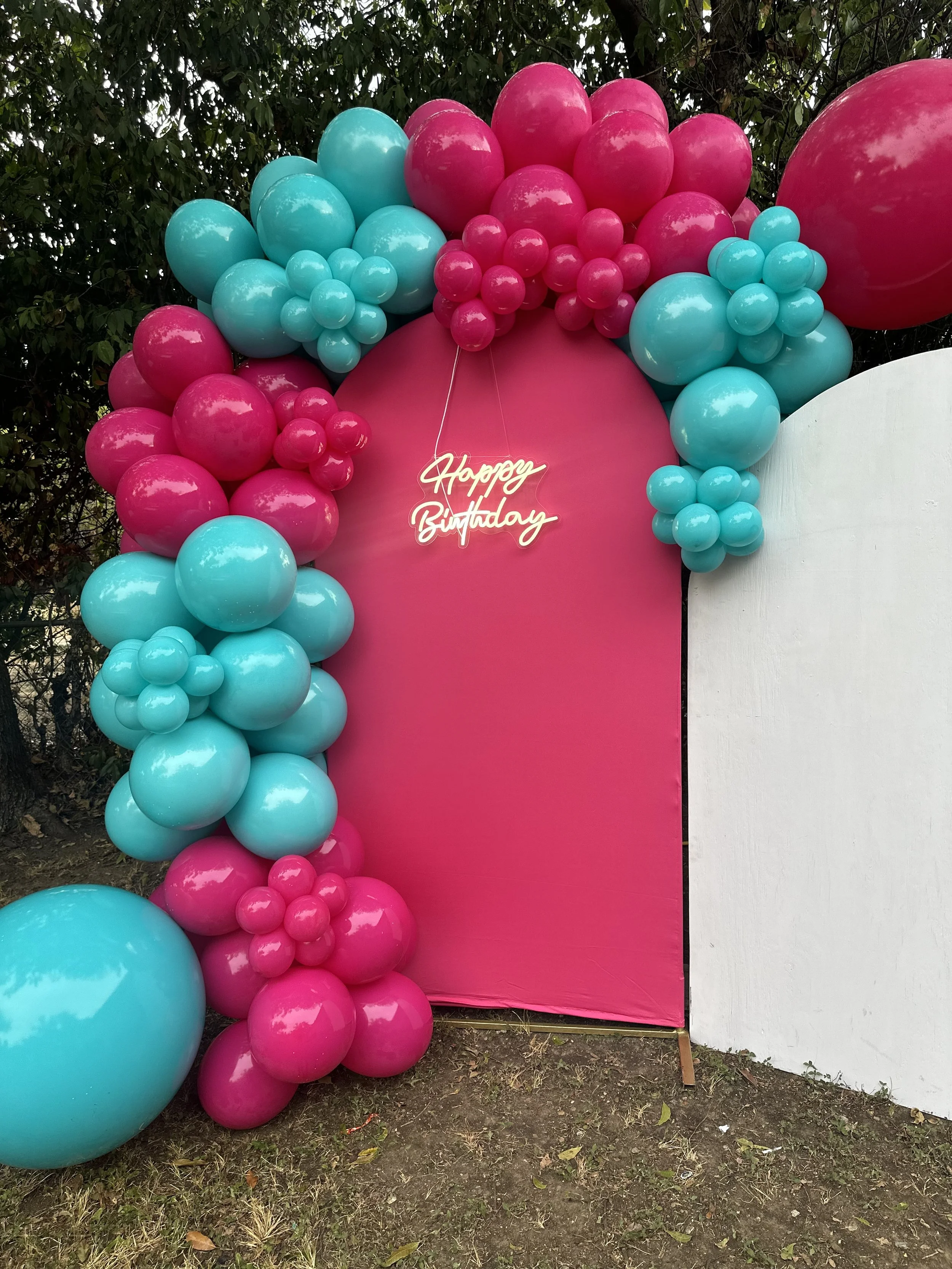 Pink and cyan balloons arranged around a pink backdrop with a white neon sign that reads 'Happy Birthday'.