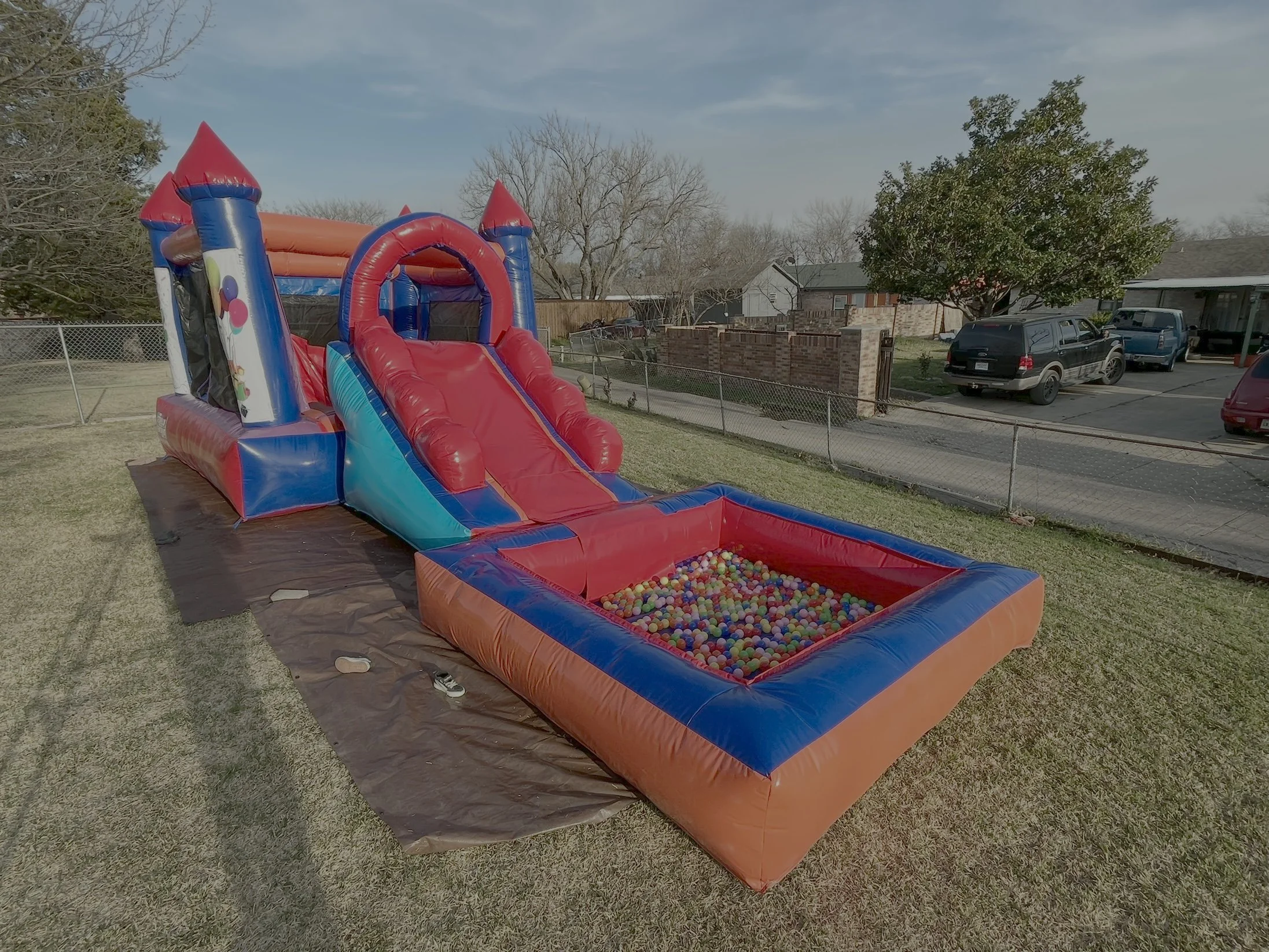 Inflatable bounce house with slide and ball pit set up in a backyard with a chain-link fence, trees, and parked cars.