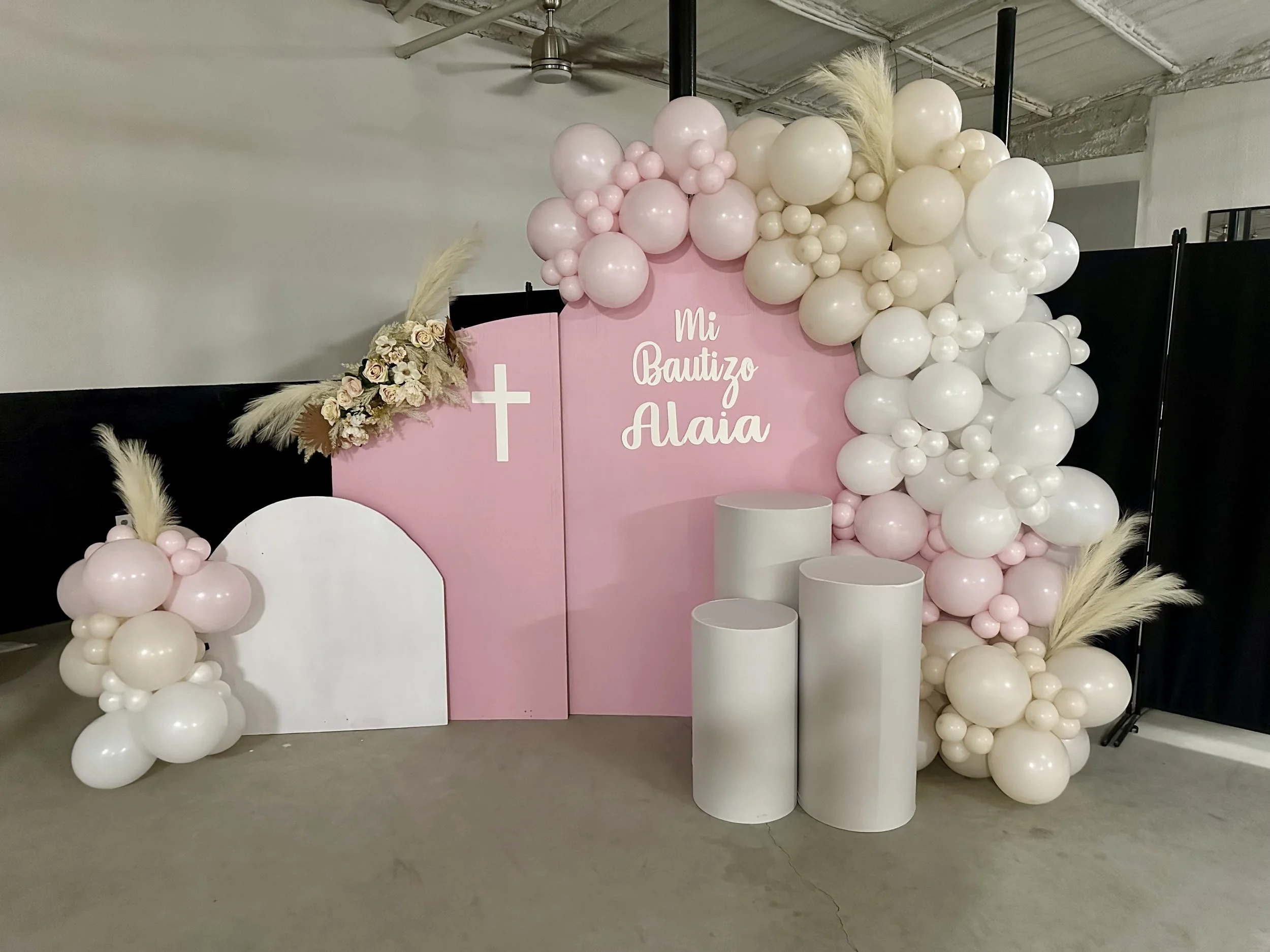 Balloon display with pink and white balloons, a pink backdrop that reads "Mi Bautizo Alaia" with a white cross, decorative floral arrangement, white pedestals, and pampas grass accents for a celebration.