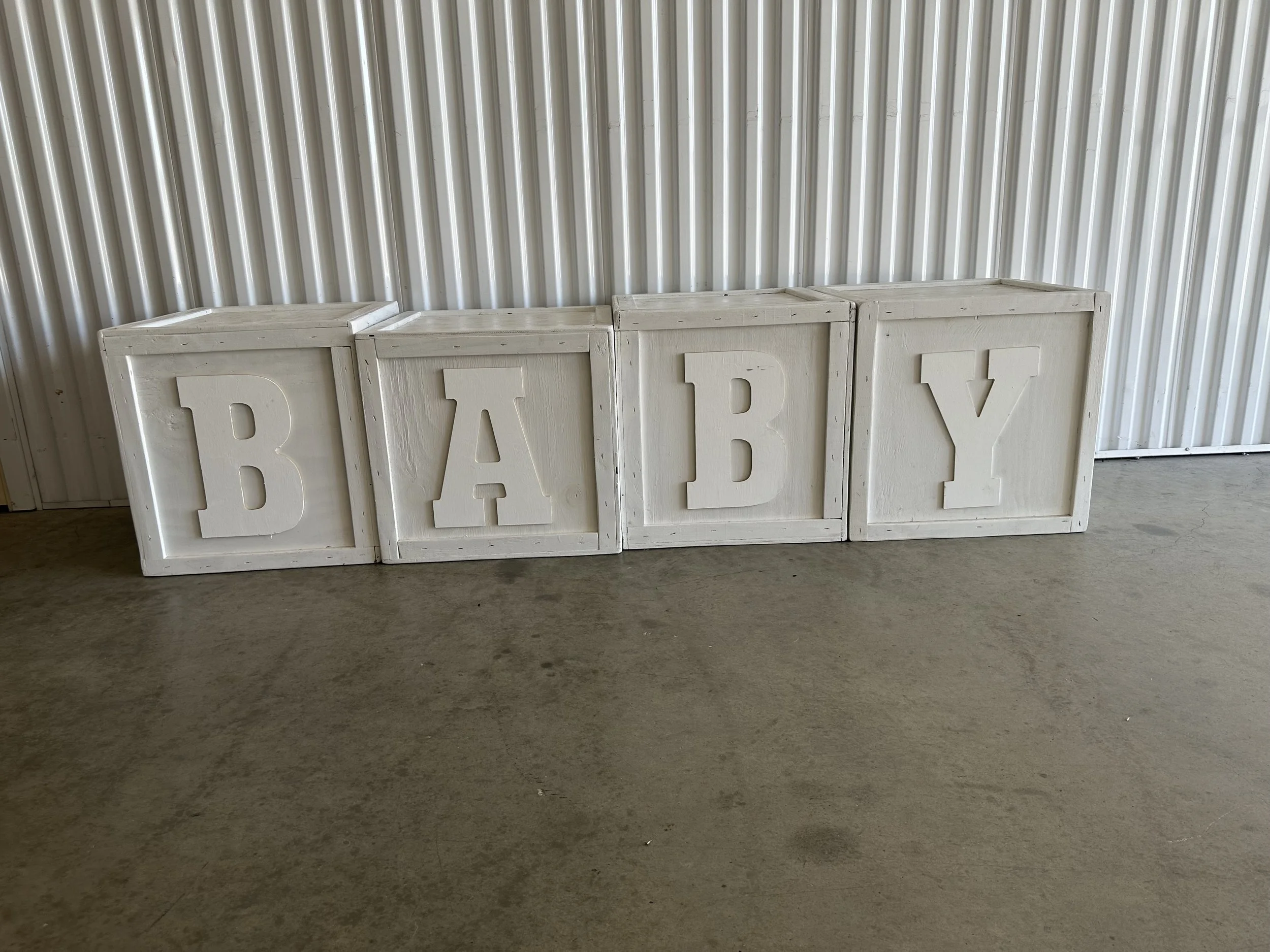 Four white blocks with large letters spelling 'BABY' are arranged against a corrugated metal wall, sitting on a concrete floor.