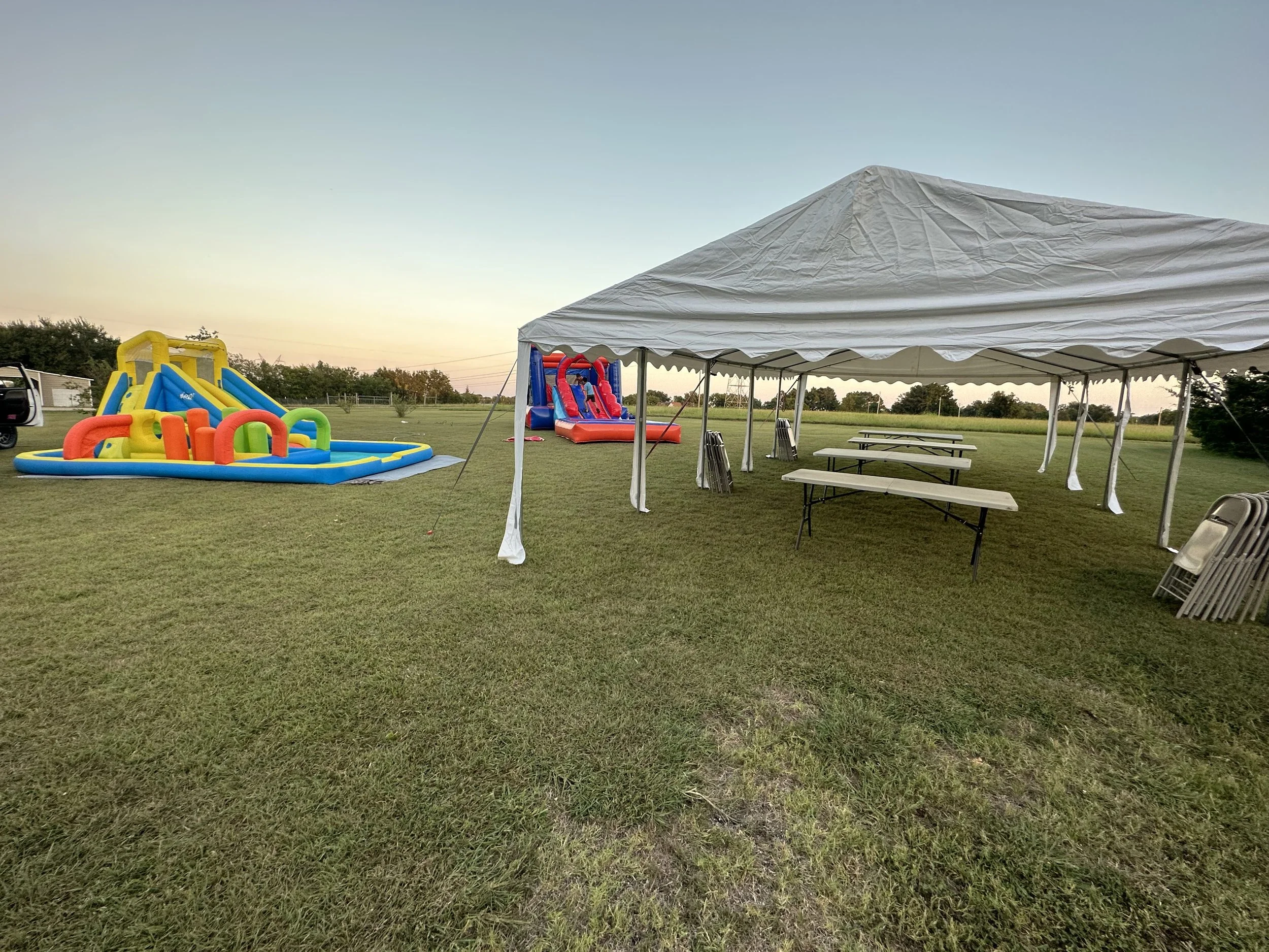 An outdoor scene at sunset with a white event tent, inflatable water slides, and folding chairs on a grassy field.
