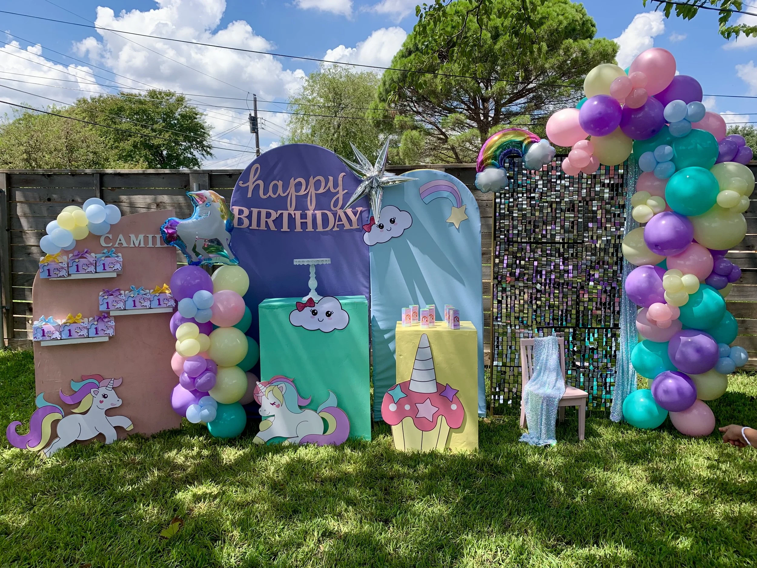 Colorful birthday party decorations featuring rainbow themed balloons, unicorn cutouts, a sign that says "happy birthday," and other whimsical decorations set against a wooden fence with trees and a partly cloudy sky in the background.