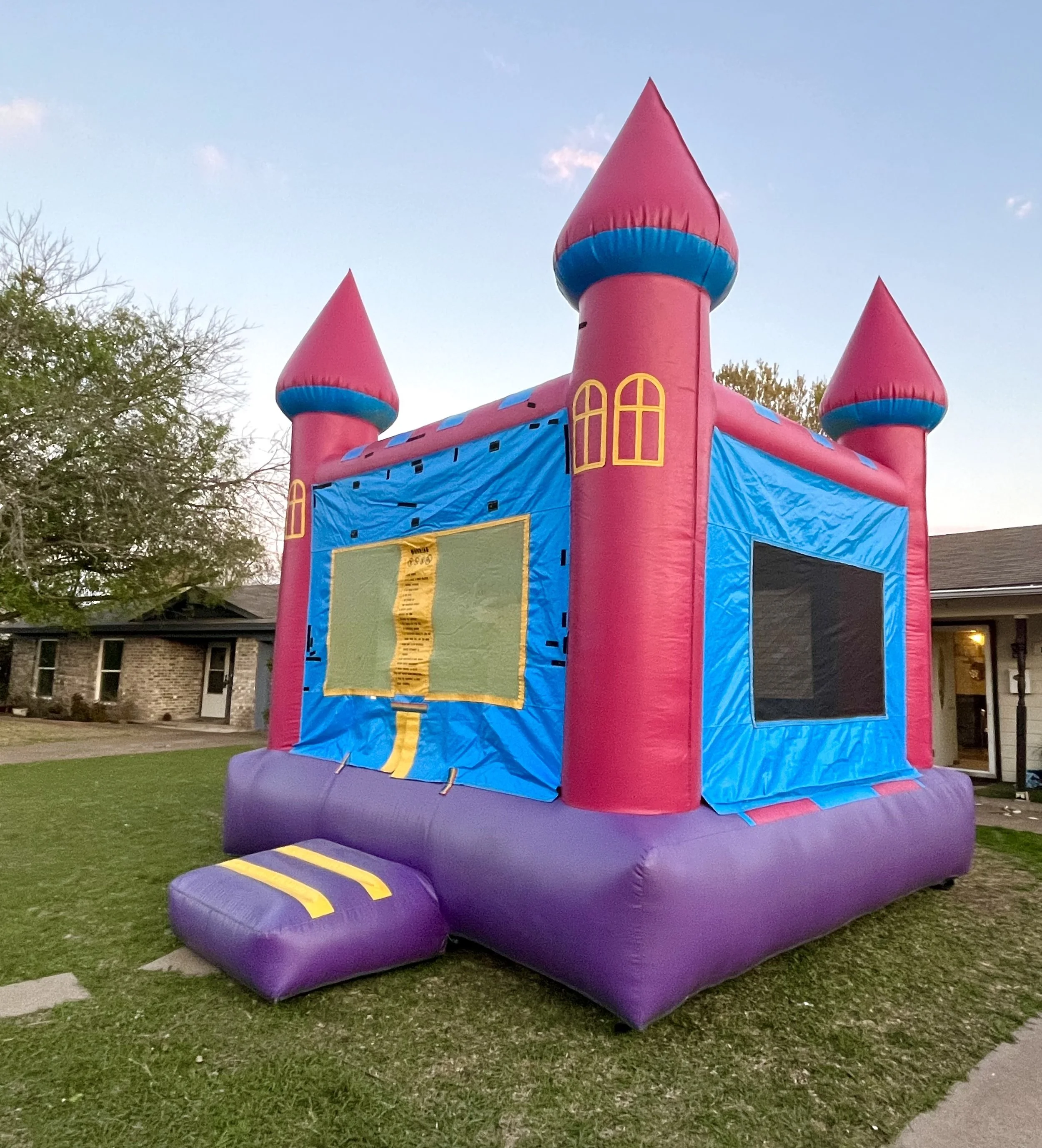 A large inflatable castle with pink towers, blue accents, and yellow window designs, situated on a grassy yard in front of houses during evening.