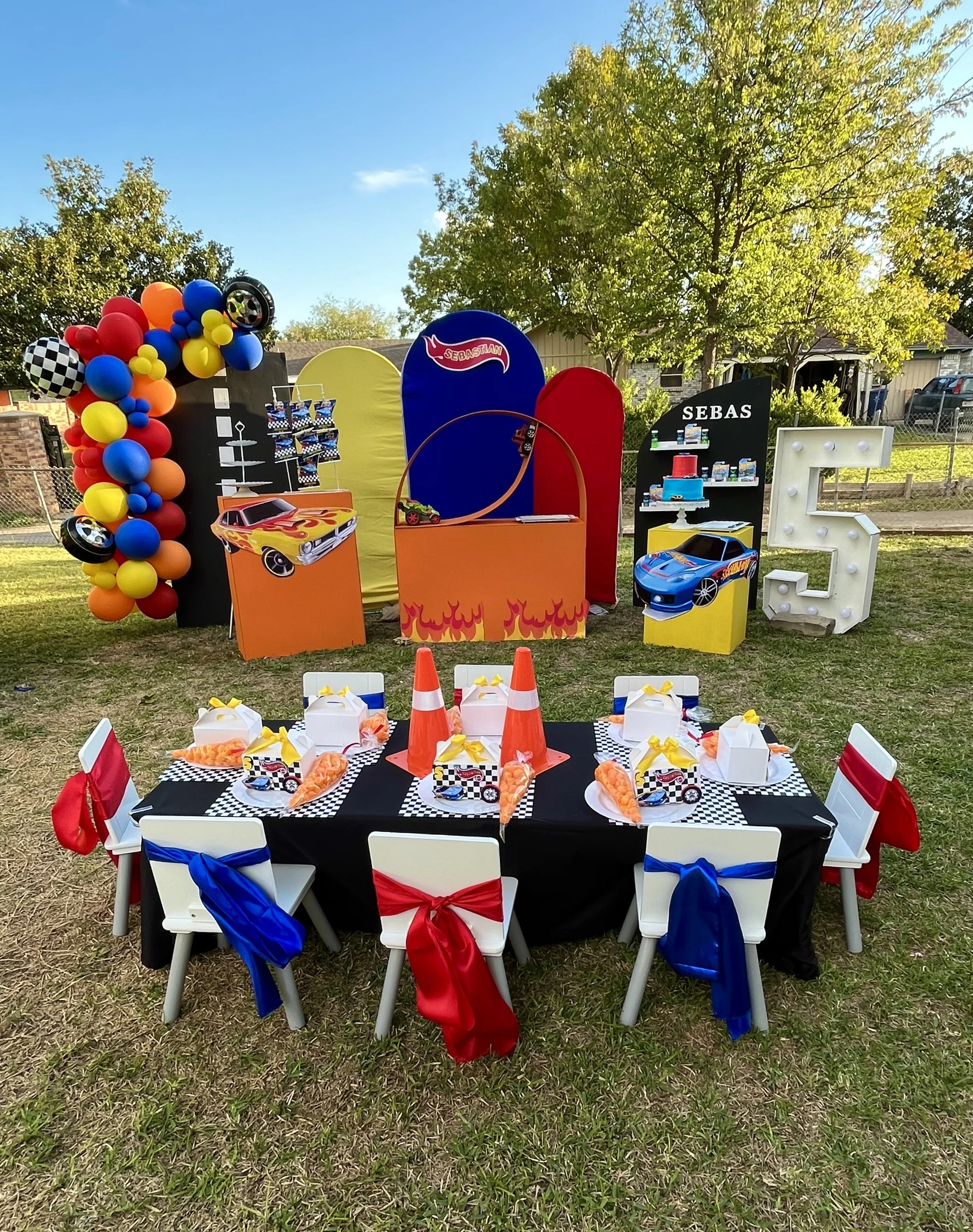 A festive outdoor birthday party setup with a race car theme. There is a decorated table with a black tablecloth, featuring race car images and orange, white, and yellow party favors. Chairs with red, blue, and white bows surround the table. In the b