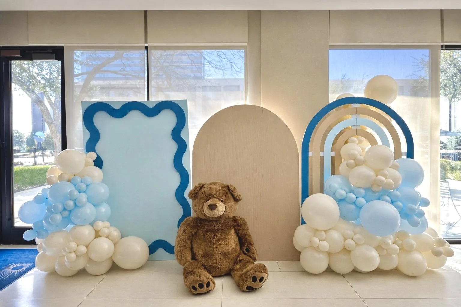 Decorative backdrop with clouds made of balloons, a teddy bear seated in front, and a blue and beige arch structure, set inside a room with large windows.