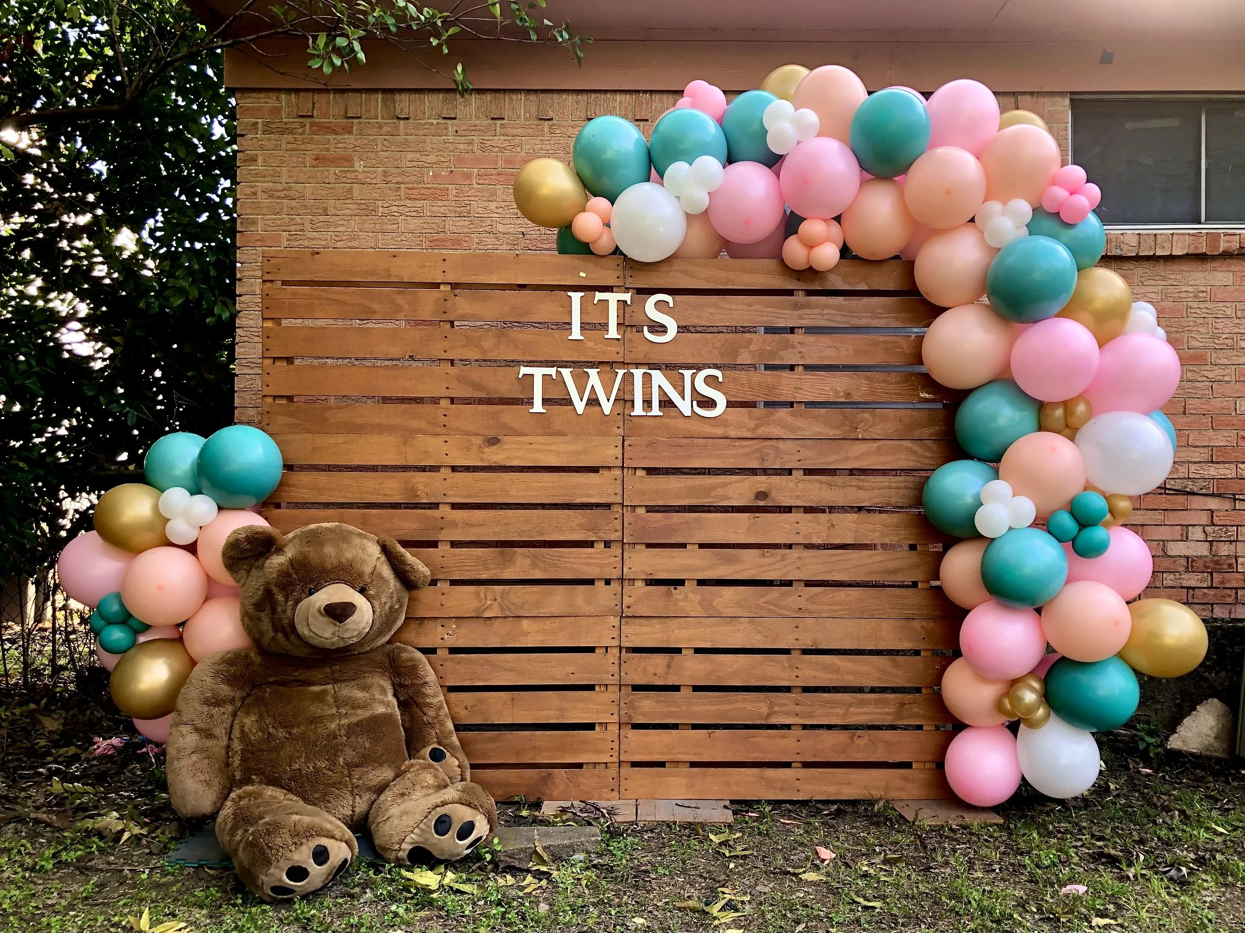 A wooden sign with the words 'IT'S TWINS' surrounded by pink, teal, white, and gold balloons. A teddy bear with glasses sits in front of the sign on the ground, partially obscuring its feet. The background features a brick wall and part of a window t