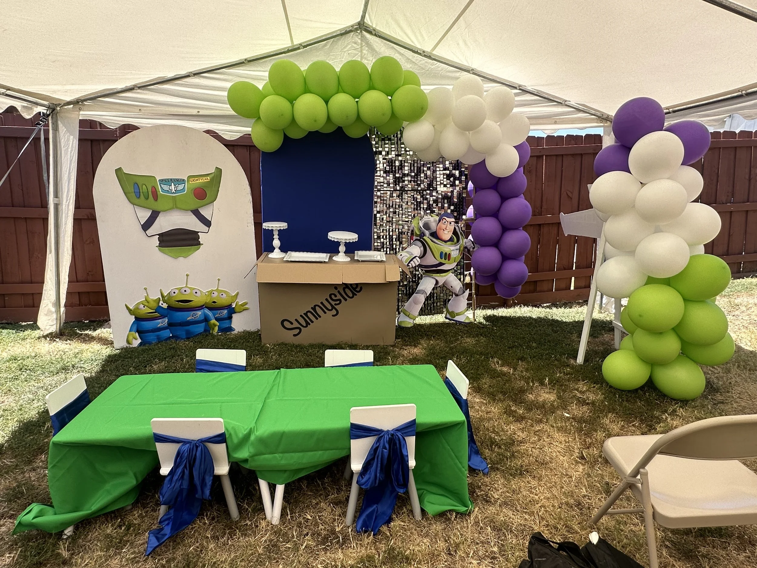 Party setup with a green and blue themed decorated table in the foreground, with chairs tied with blue bows, under a white tent. In the background, balloons in green, white, and purple, along with a 'Toy Story' themed standee featuring Buzz Lightyear