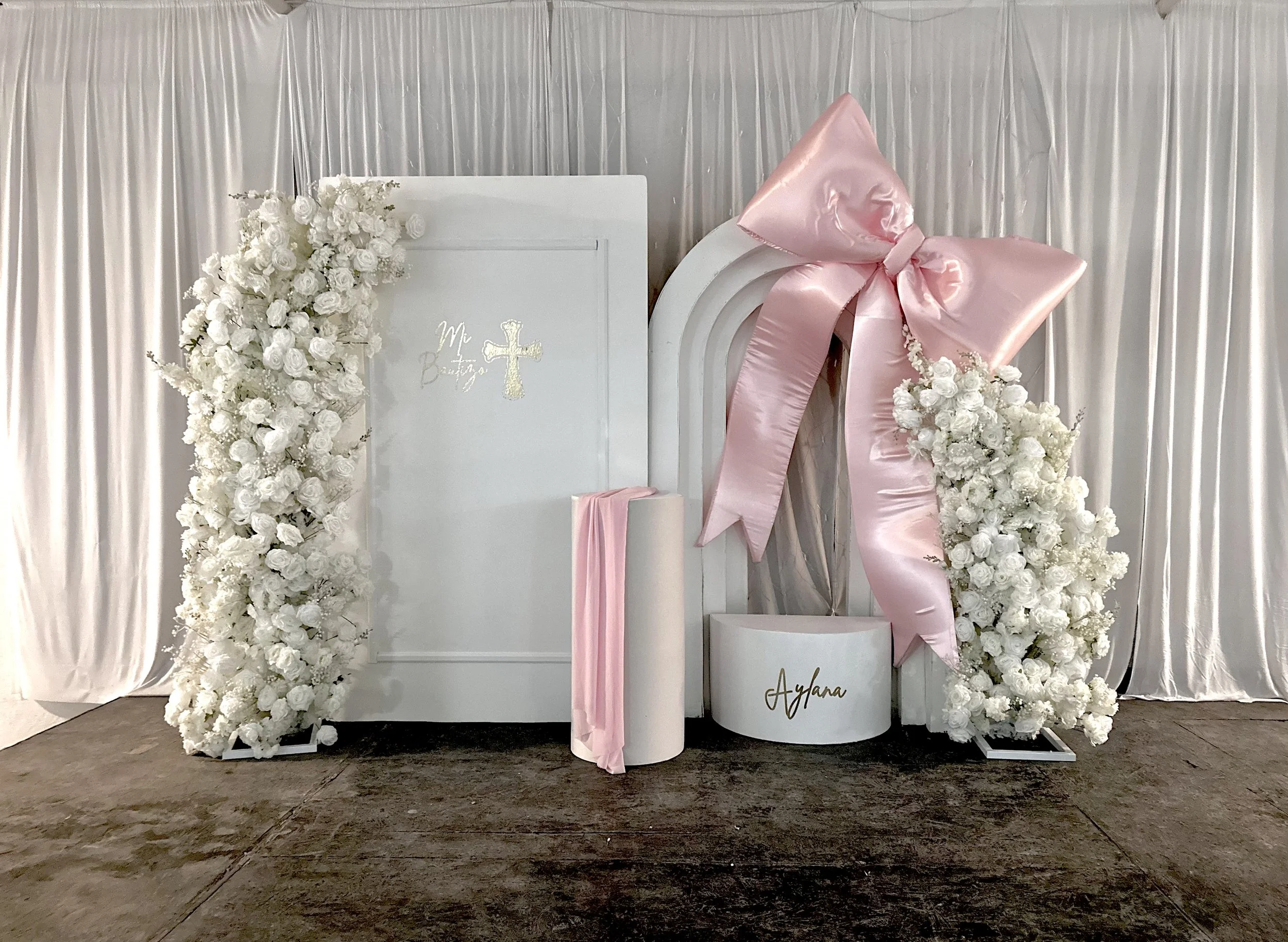 A decorated wedding or celebration backdrop with white flowers, pink ribbons, a white arch, a white box with gold text, and a backdrop of white curtains.