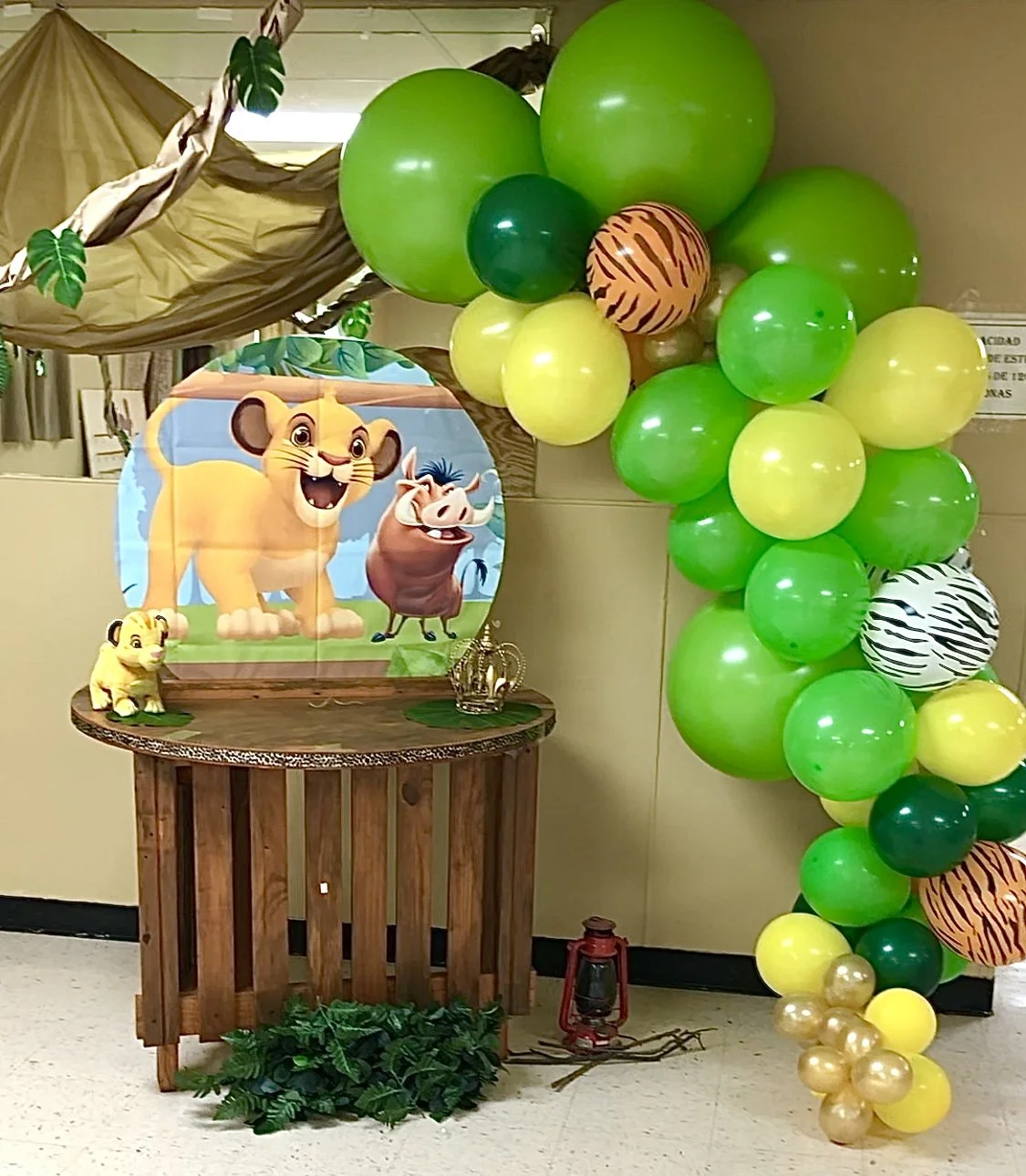Jungle themed birthday party decoration with green, yellow, gold, black and white balloons, a wooden table, a backdrop featuring The Lion King characters, a small lion figure, a zebra print balloon, and jungle leaves and a lantern as decor elements.
