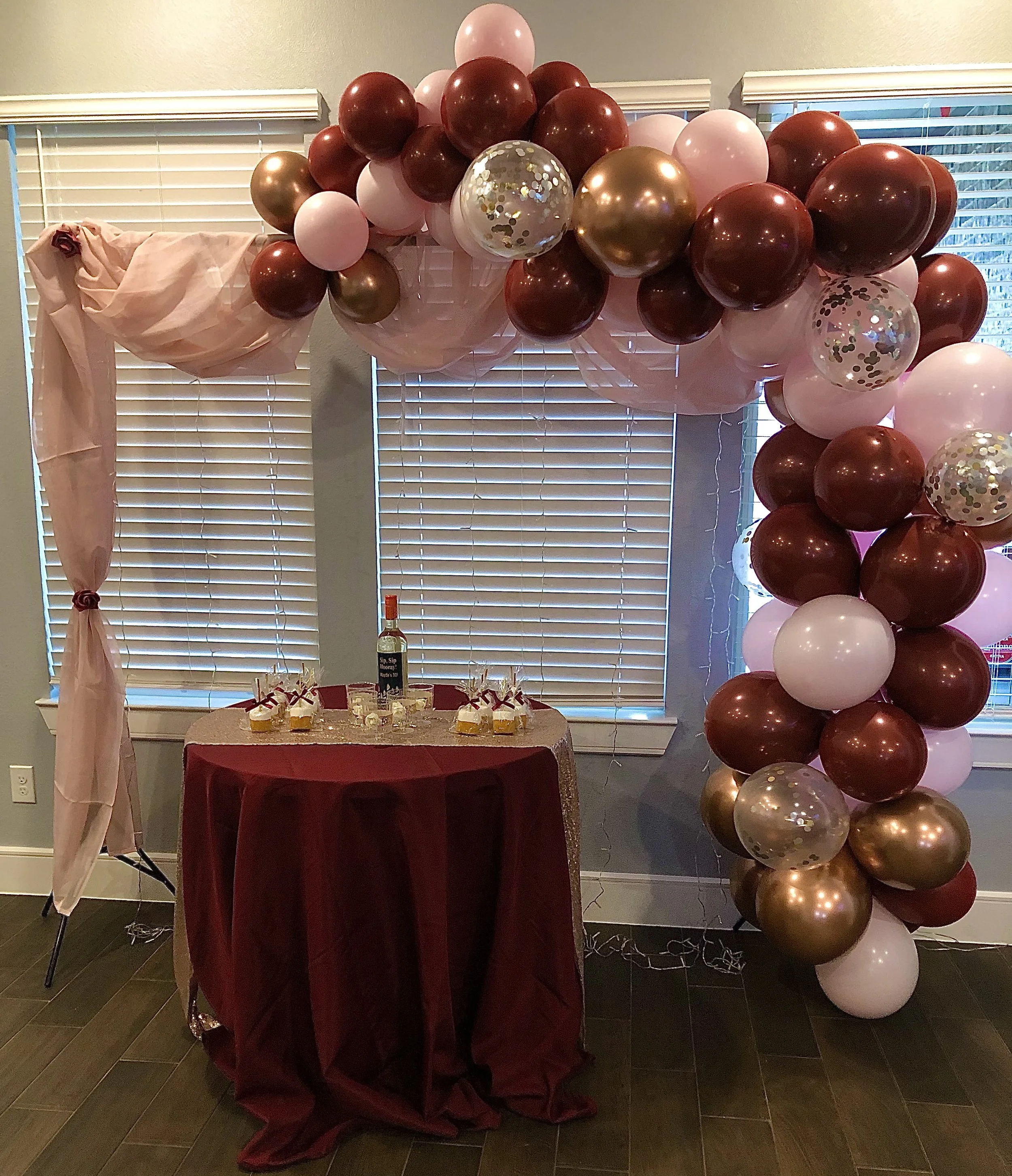 A balloon decoration arrangement with pink, white, gold, and dark red balloons, placed above a table draped with dark red fabric, in front of window blinds.