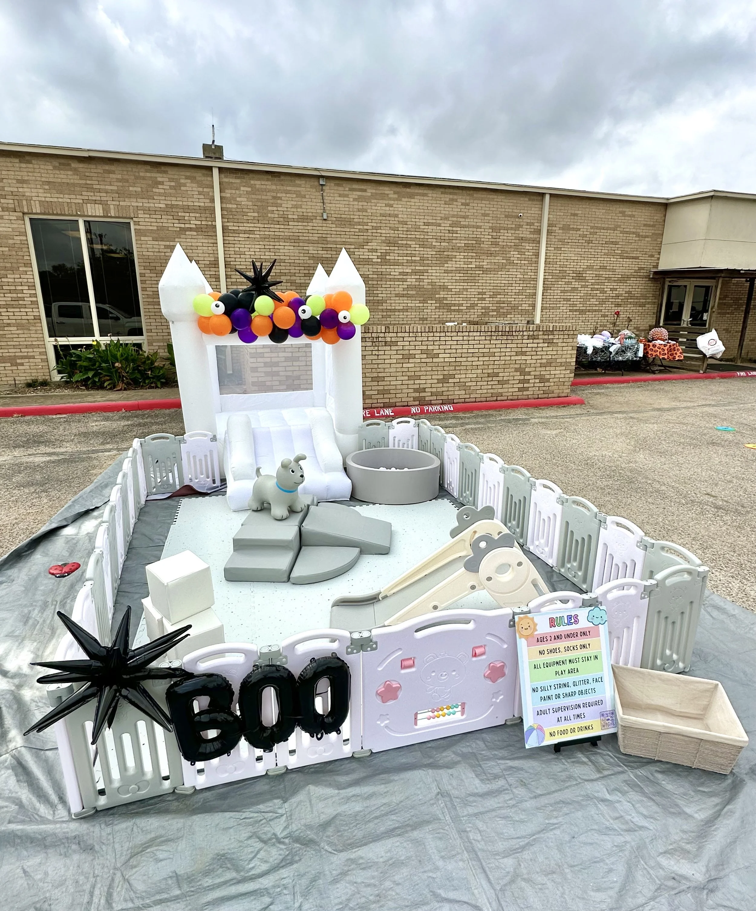 Outdoor event setup with a white inflatable bounce house decorated with colorful balloons resembling a witch's hat and a black star, a small gray dog figure, black spider props, and a folding table with various items in the background. A sign lists r