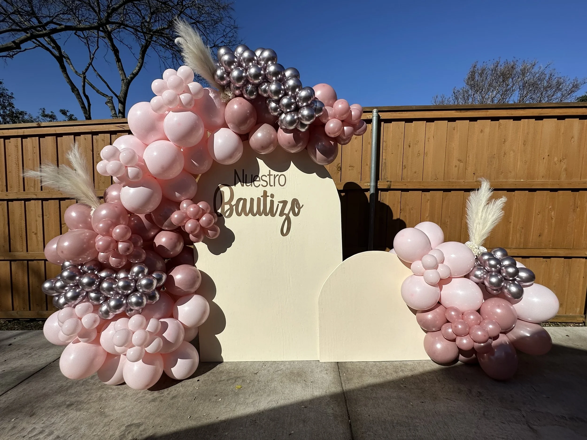 Balloon display with pink and silver balloons arranged in an arch and around a white sign that reads 'Nuestro Bautizo' against a wooden fence and clear blue sky.