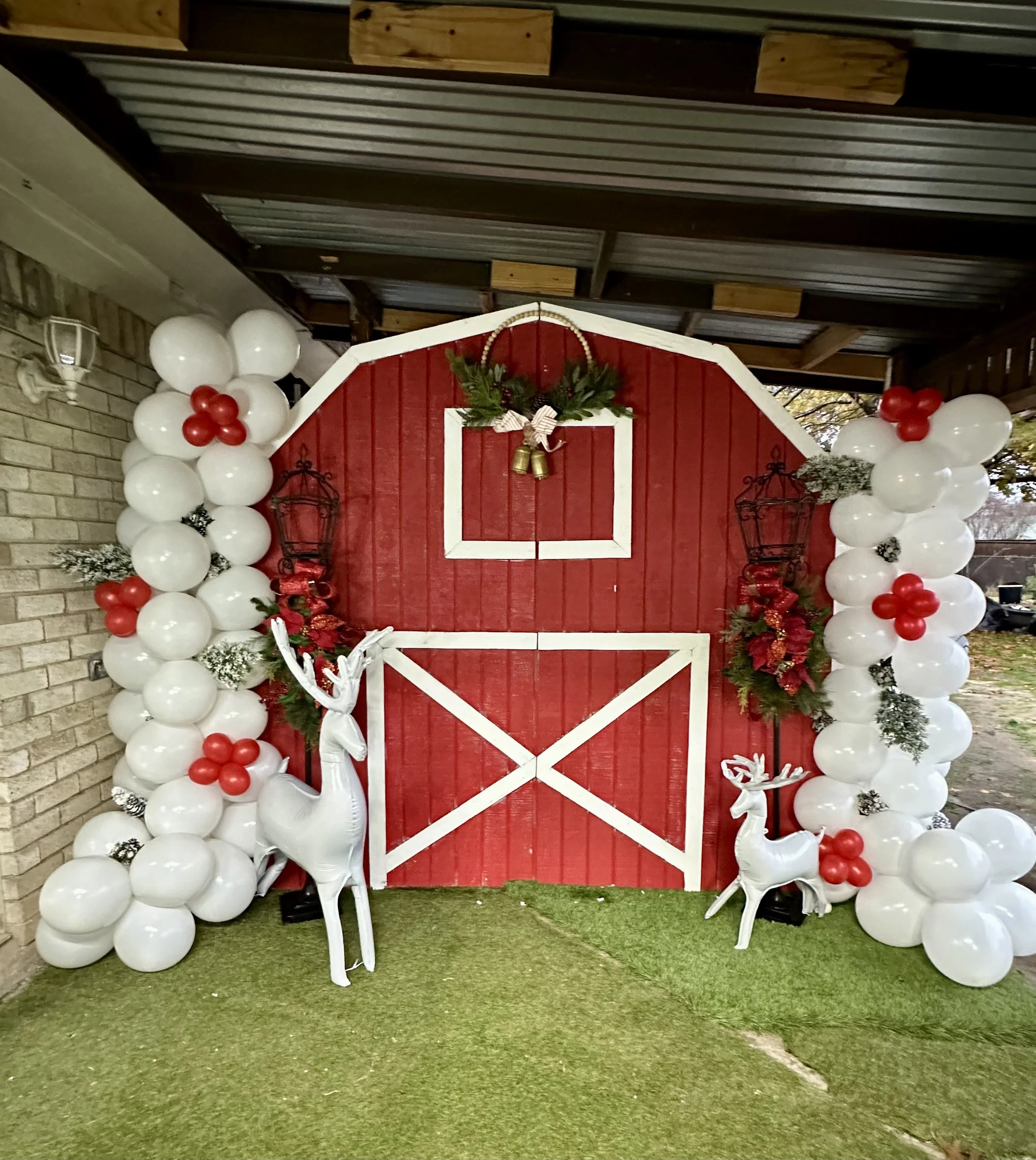 Decorative red barn backdrop framed by white and red balloon arch with Christmas greenery, ornaments, and white reindeer statues, set on artificial grass.