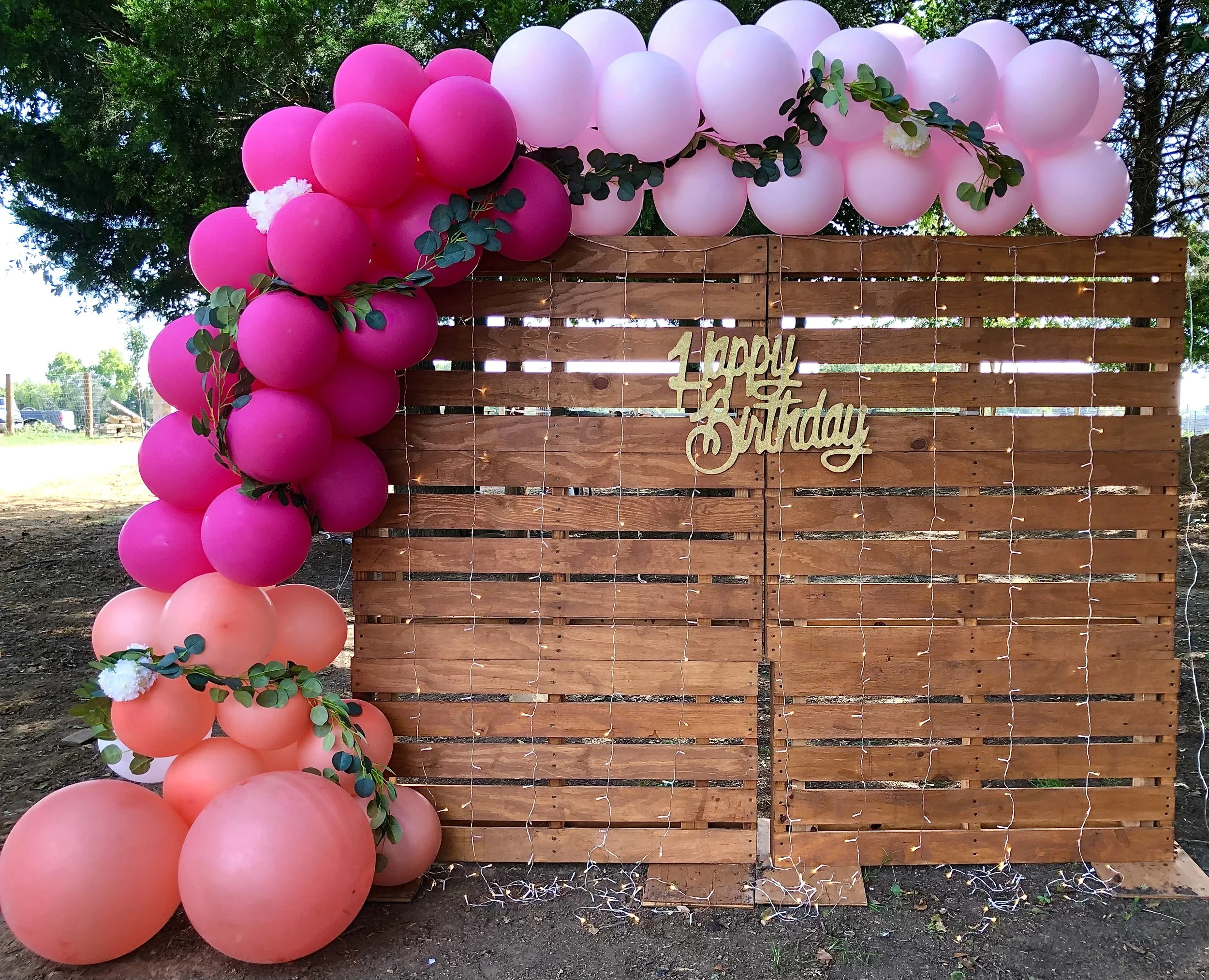 A wooden backdrop decorated with pink and peach balloons and green leaves. The backdrop features a sign that says 'Happy Birthday' with string lights draped across.