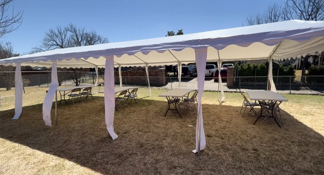 White event tent set up on a grassy yard with tables and chairs inside, surrounded by a chain-link fence and parked cars in the background on a sunny day.