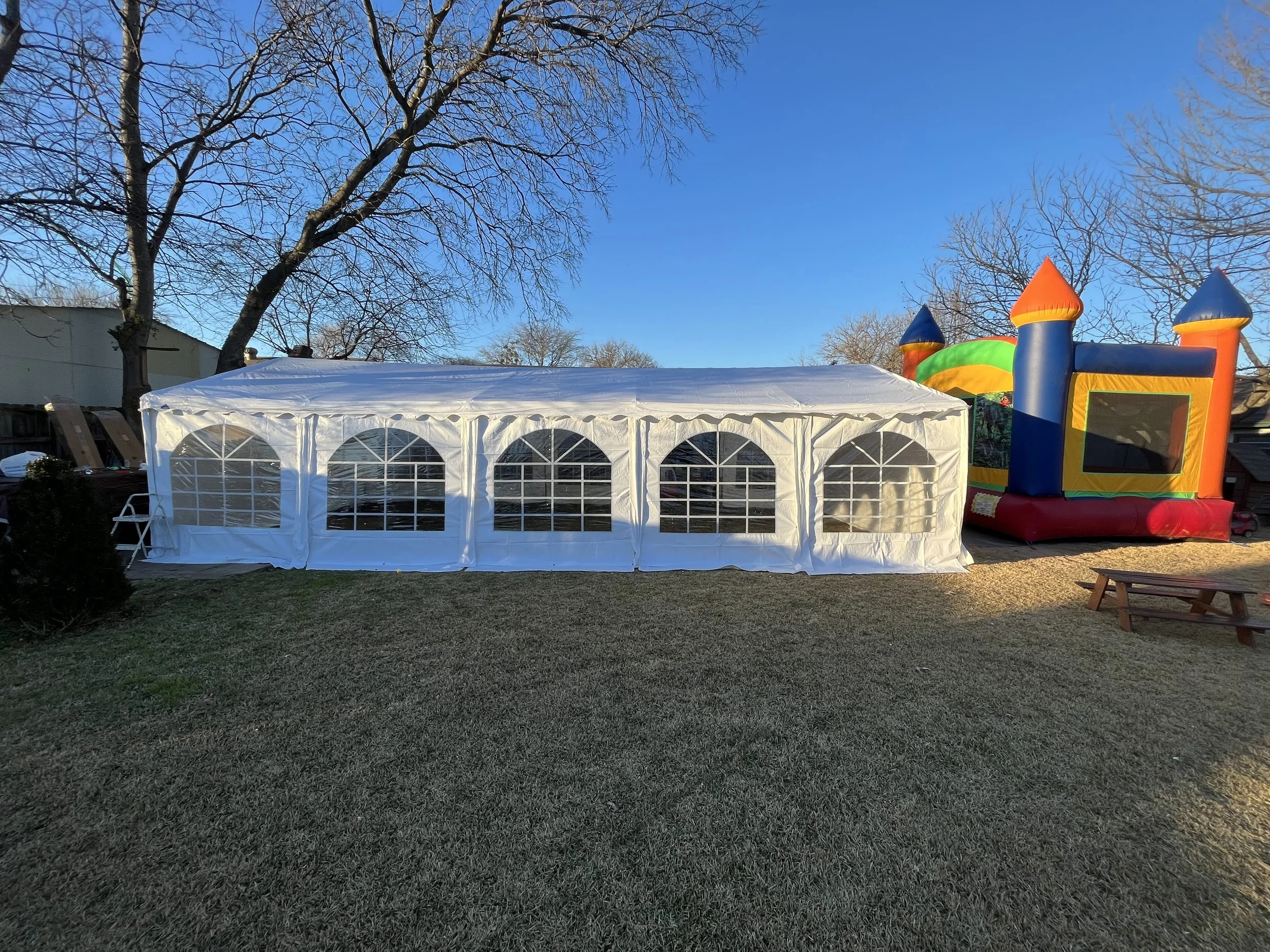 An outdoor scene with a large white event tent on green grass, tall bare trees, a colorful inflatable bounce house on the right, and clear blue sky.