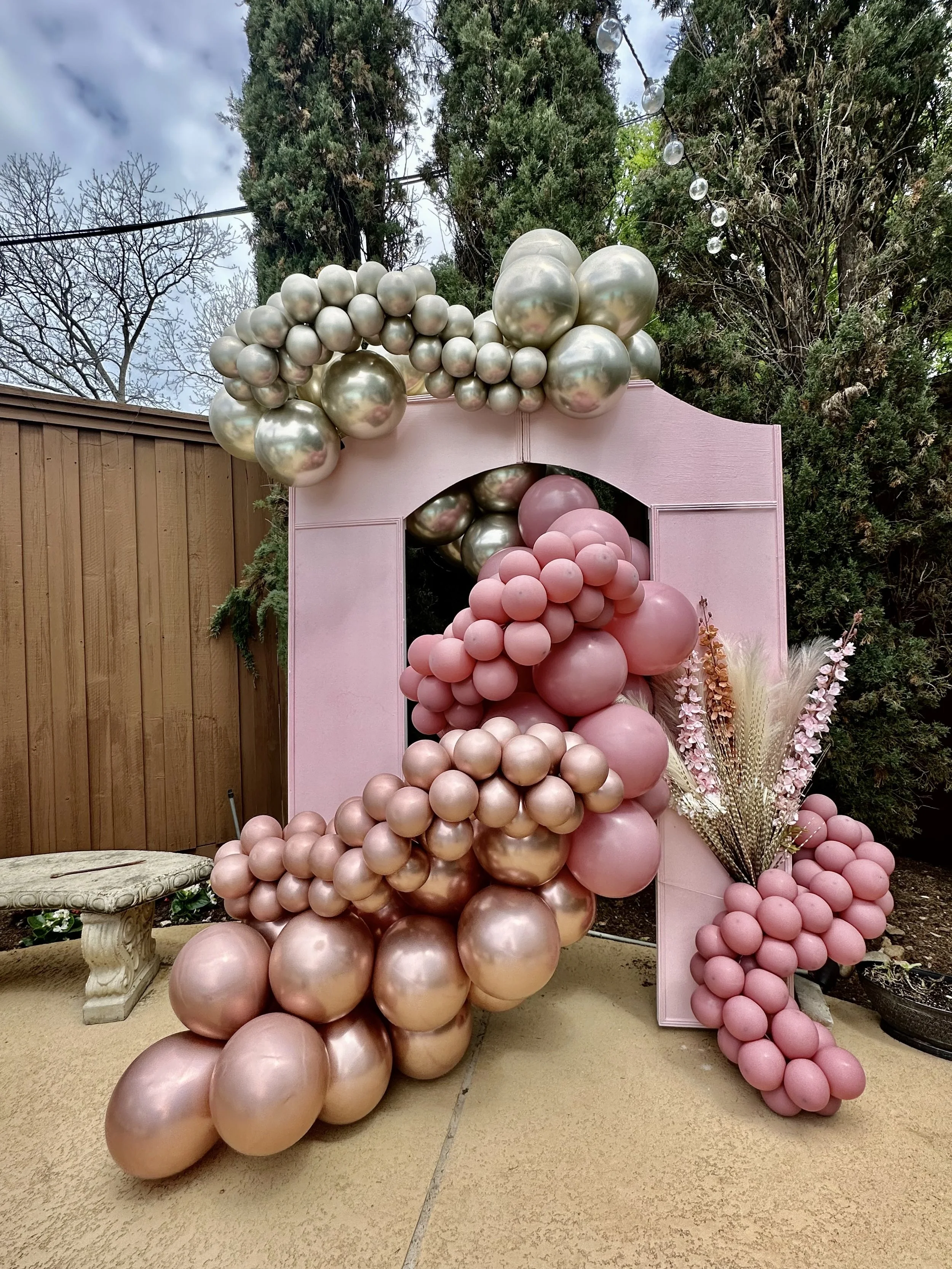 Decorative background with pink and gold balloons arranged around a pink frame, with pink balloons cascading down to the ground and a bouquet of dried flowers on the right side.